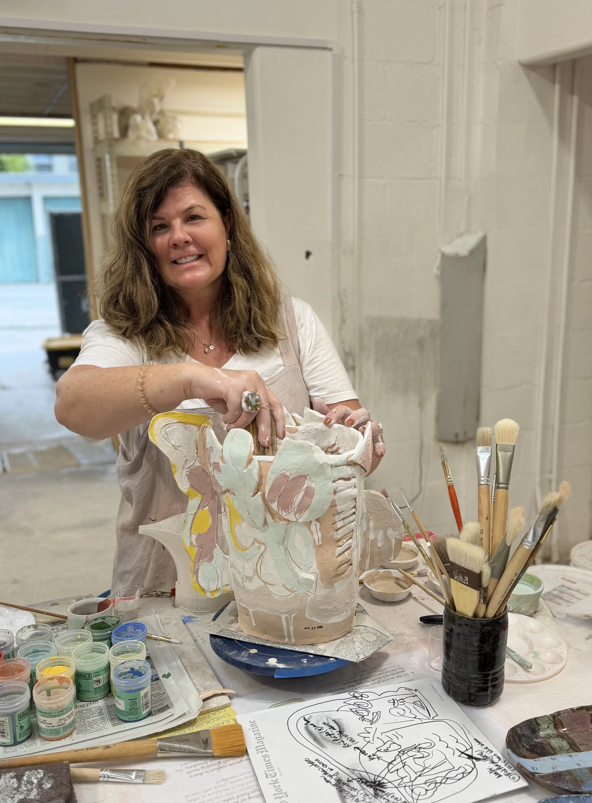 A woman is holding a decorative ceramic vase she is painting, with paint supplies and brushes on the table in front of her. The workspace has art supplies and a partly painted vase.