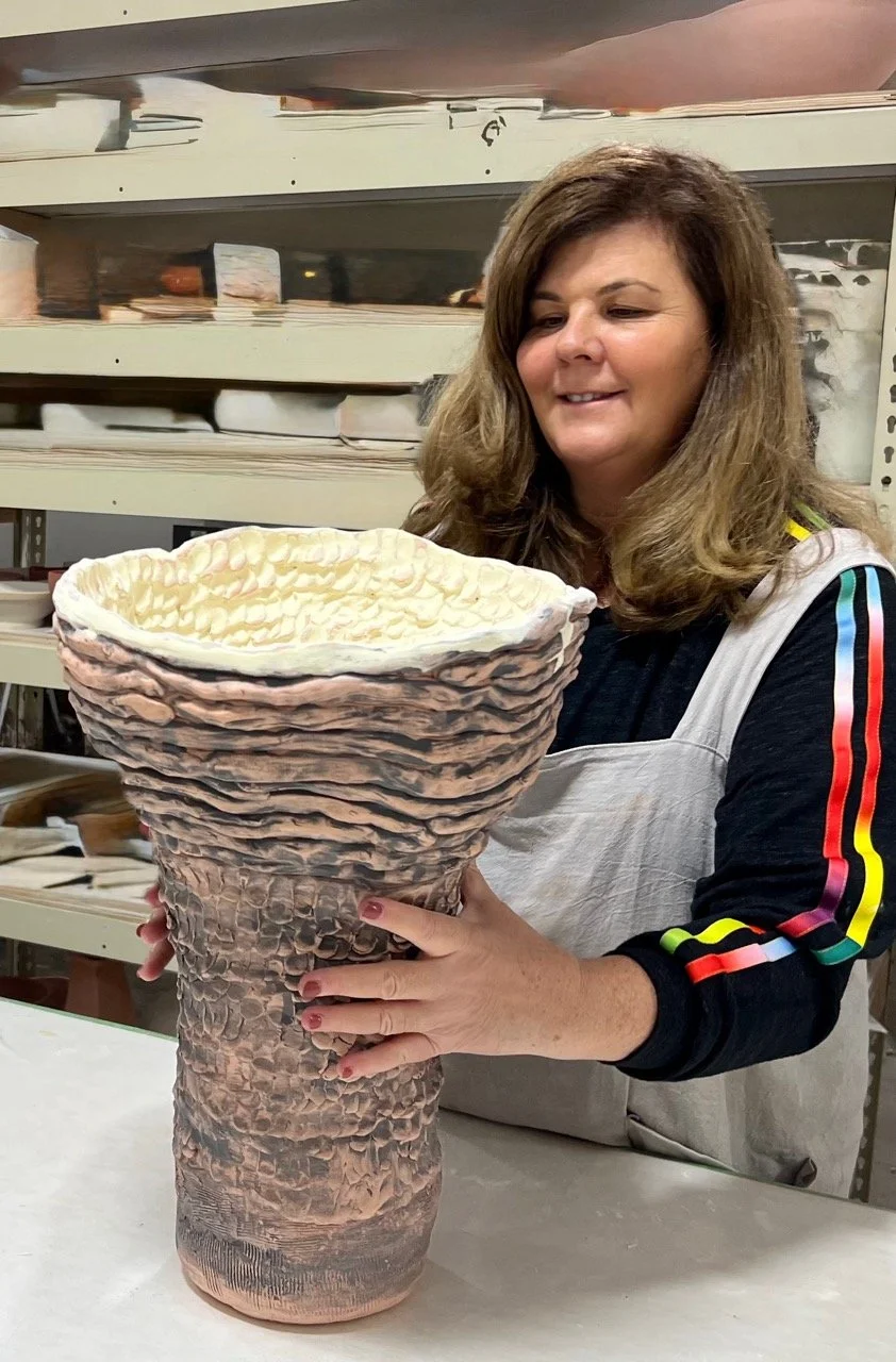 A woman with brown hair wearing a beige apron and a black shirt with colorful stripes holds a large, ceramic vessel resembling a textured, outdoor-borne plant pot, with a yellow interior surface, inside a workshop or studio.