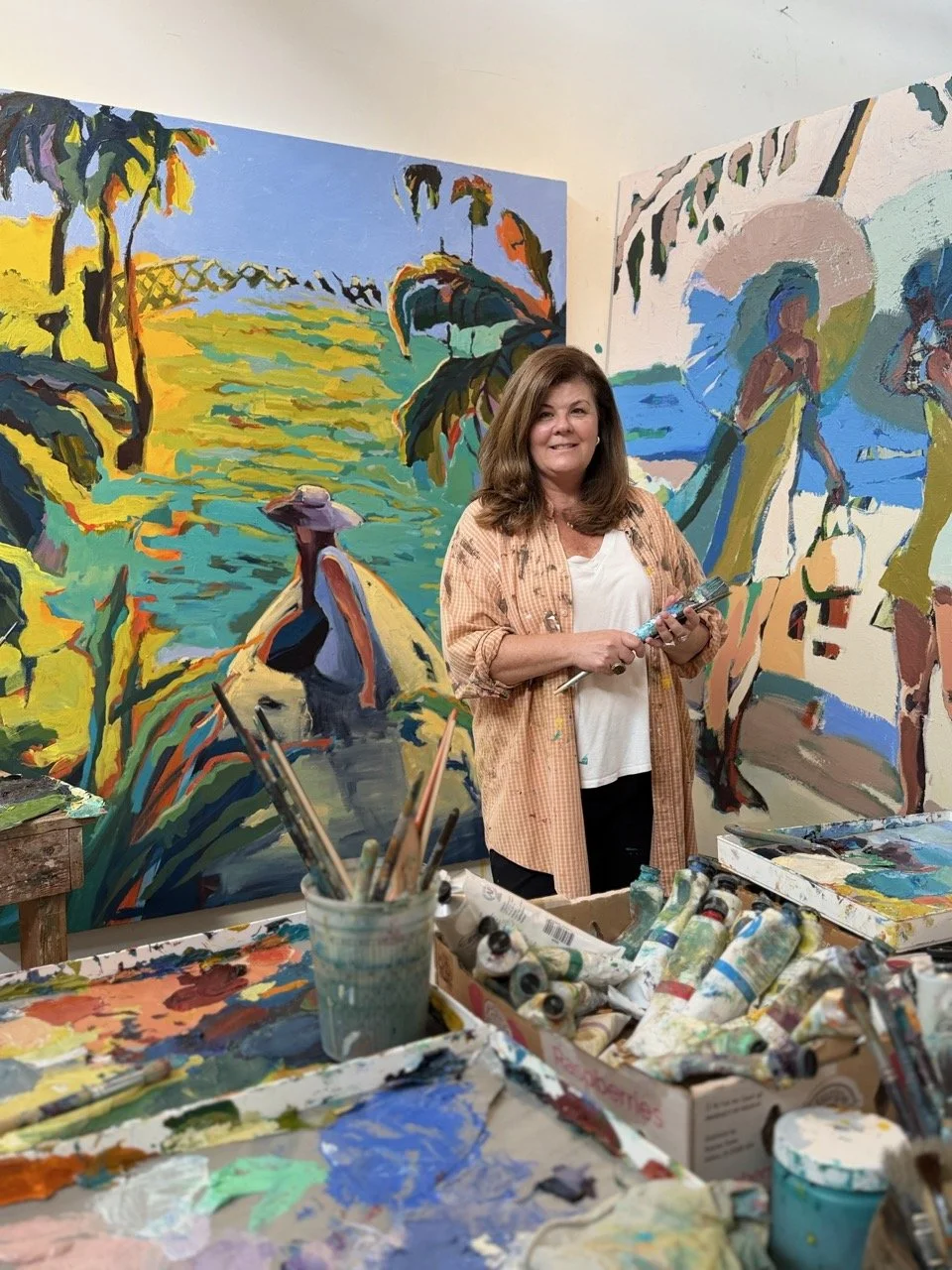 A woman standing in front of colorful landscape paintings in an art studio, surrounded by paint tubes and brushes.