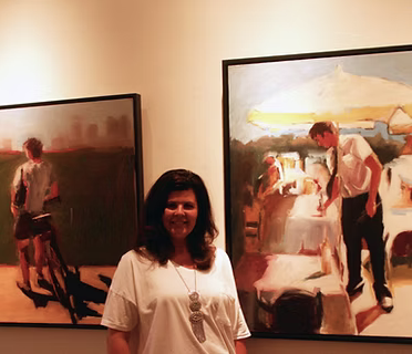 A woman standing in front of two abstract paintings of people at a beach or seaside setting.