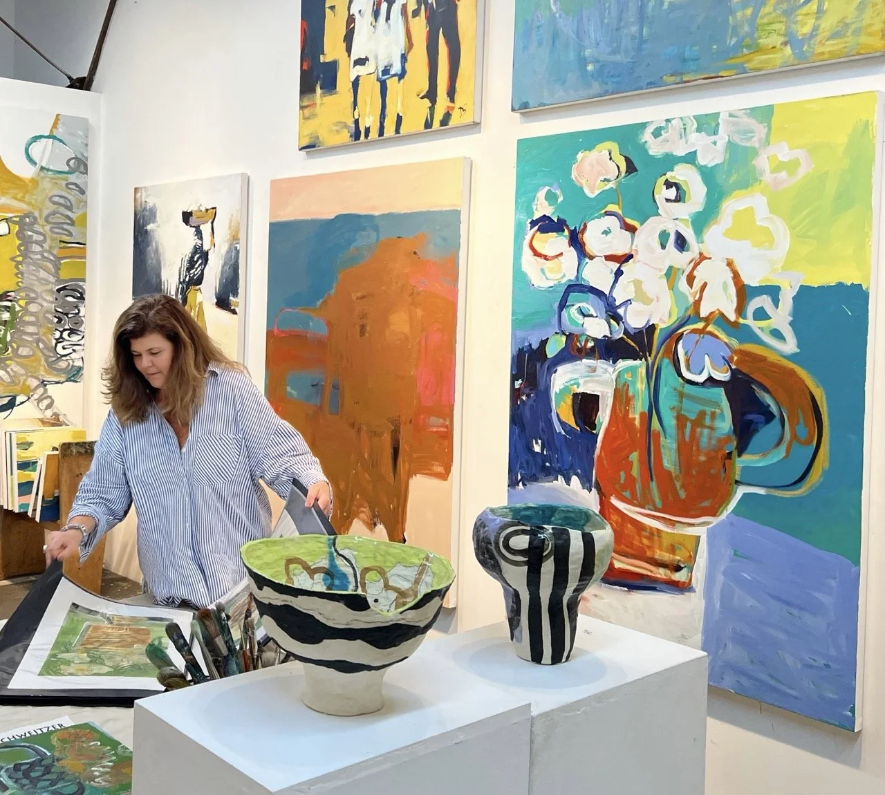 An art gallery with a woman examining artwork at a table. Various colorful paintings on the walls and ceramic vases on display.