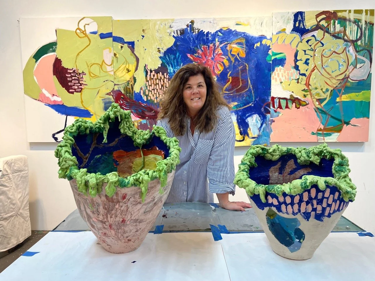 Woman with wavy brown hair smiling in art studio, standing behind large, colorful ceramic pots with green, textured rims, in front of an abstract painting with bright colors and bold brushstrokes.