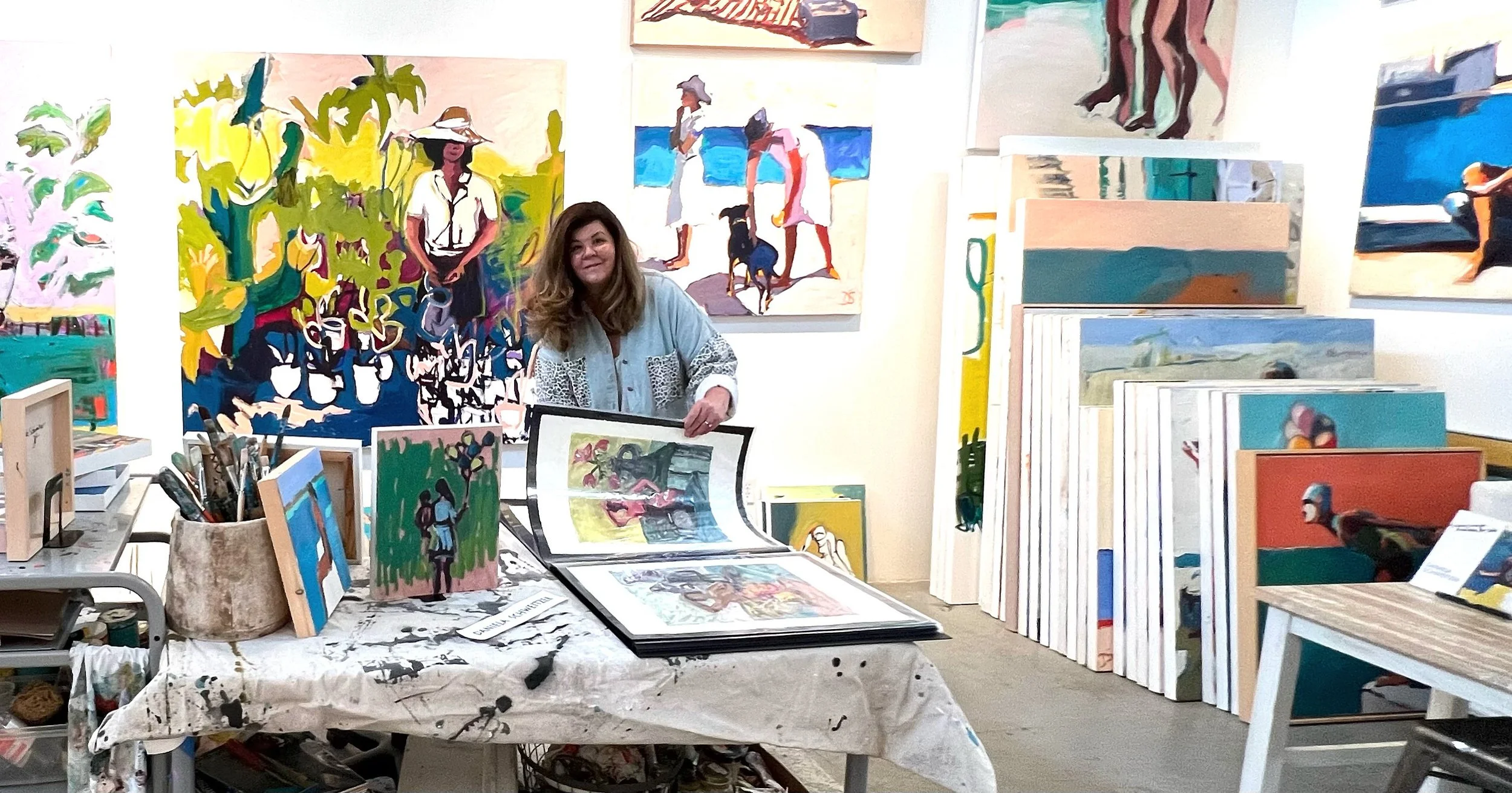 An artist woman sitting at a table in an art studio, surrounded by colorful paintings and art supplies, with more paintings on the walls and stacked around.