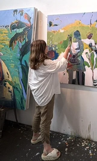 A woman in a white shirt and beige pants is painting on a large canvas in an art studio, with another large canvas leaning against the wall nearby.