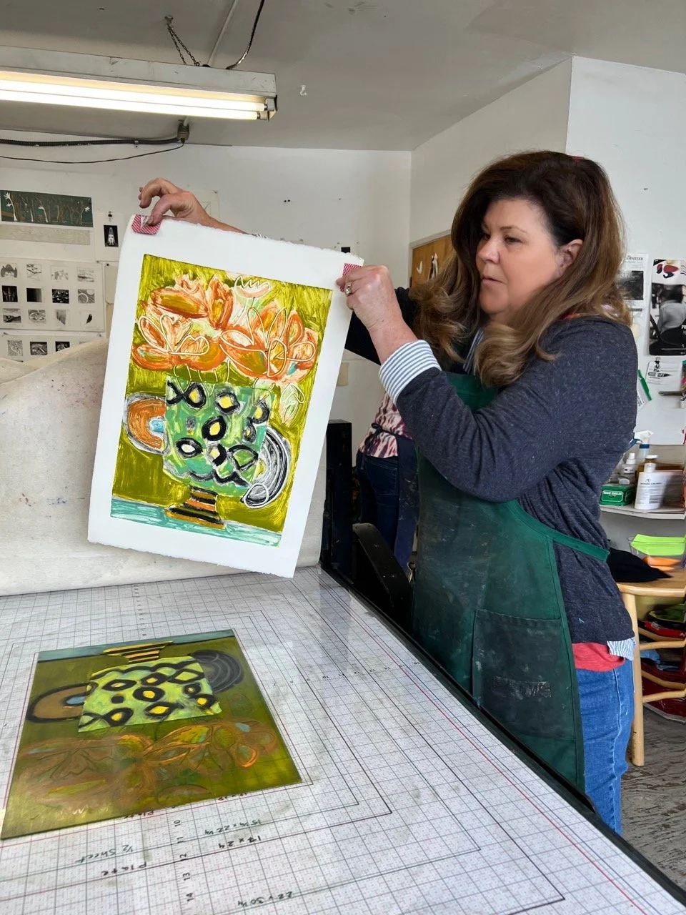 Woman in a green apron holding up a colorful abstract painting of a vase with flowers, in an art studio.