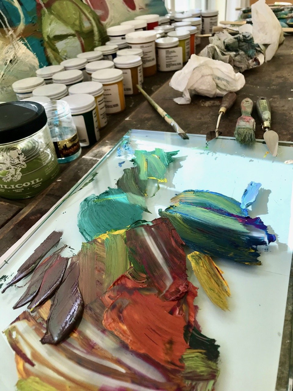 An artist's workspace with a palette loaded with colorful oil paints, several jars of paint on a wooden table, paintbrushes, and painting supplies in the background.