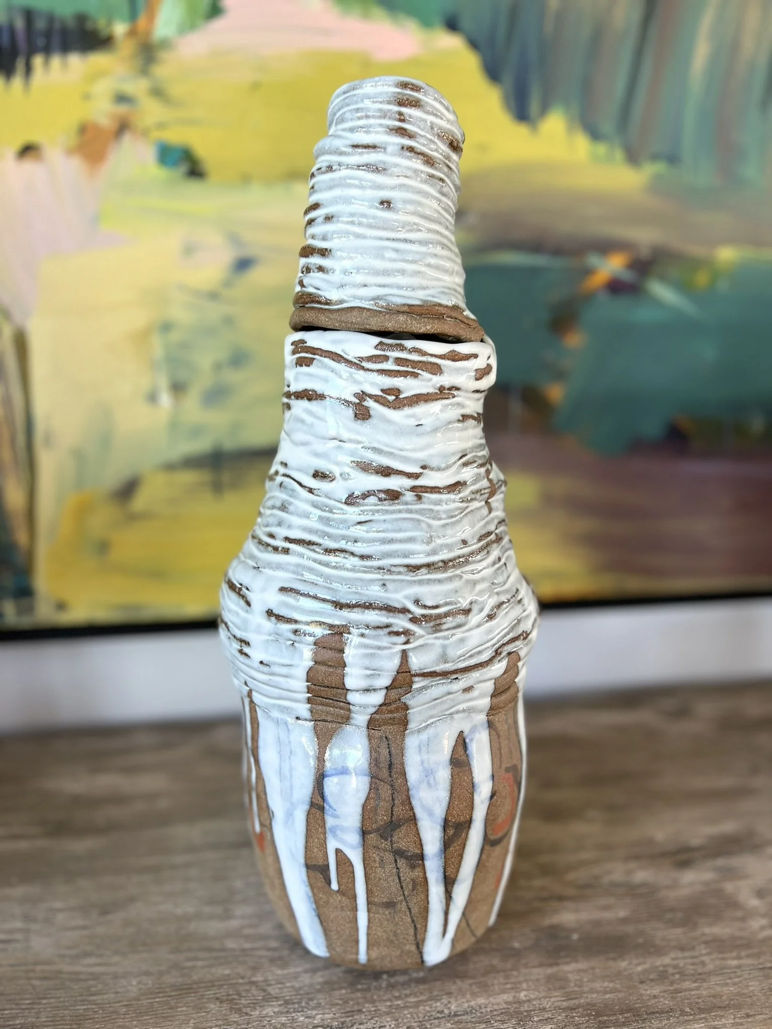 A ceramic cone-shaped sculpture with a textured, layered surface painted with white and brown glaze, sitting on a wooden surface against an abstract, colorful background.