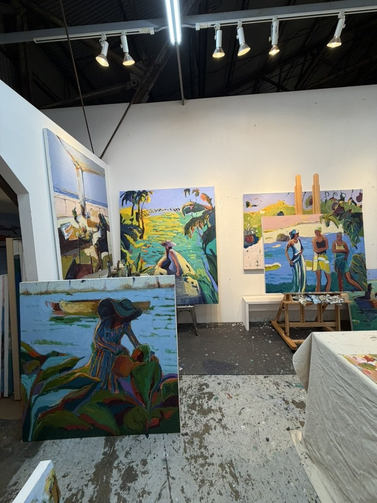 Multiple colorful landscape paintings in progress, depicting scenes of people by the water, on an art studio wall.