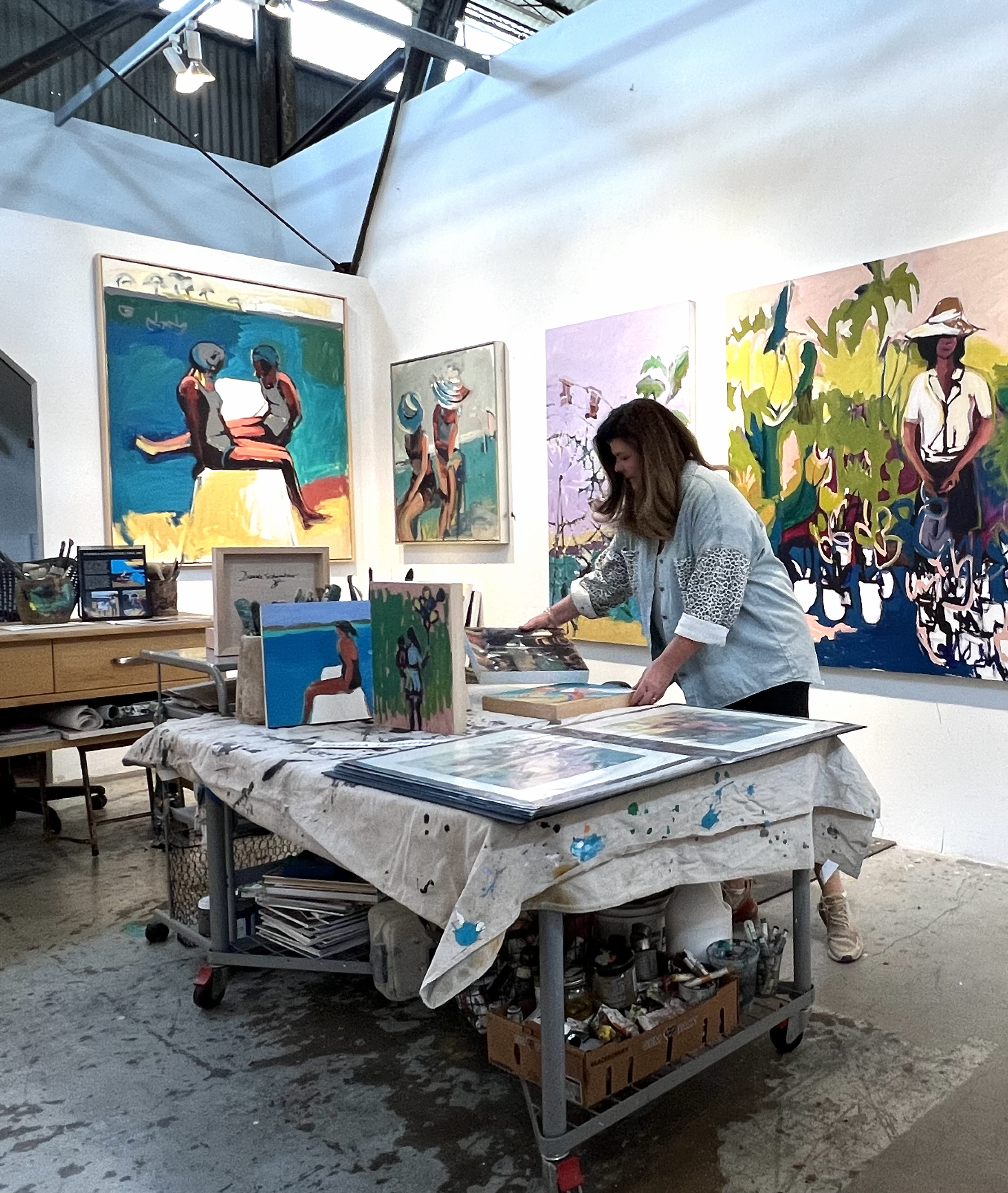 An art gallery displaying colorful paintings of people and nature, with a woman arranging artwork on a table in the foreground, and a display of multiple framed art pieces on a table and on the wall.
