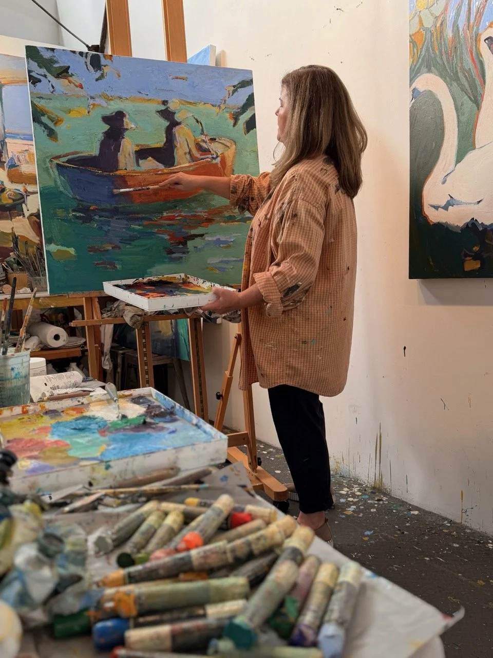 An artist in a studio painting an impressionistic landscape scene of two people in a boat on a body of water, surrounded by paintings, art supplies, and paint tubes.