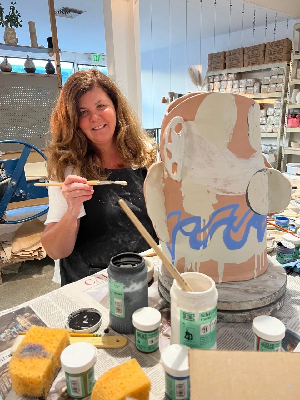 A woman painting a large ceramic pot in an art studio, surrounded by painting supplies and shelves filled with other ceramic pieces and supplies.