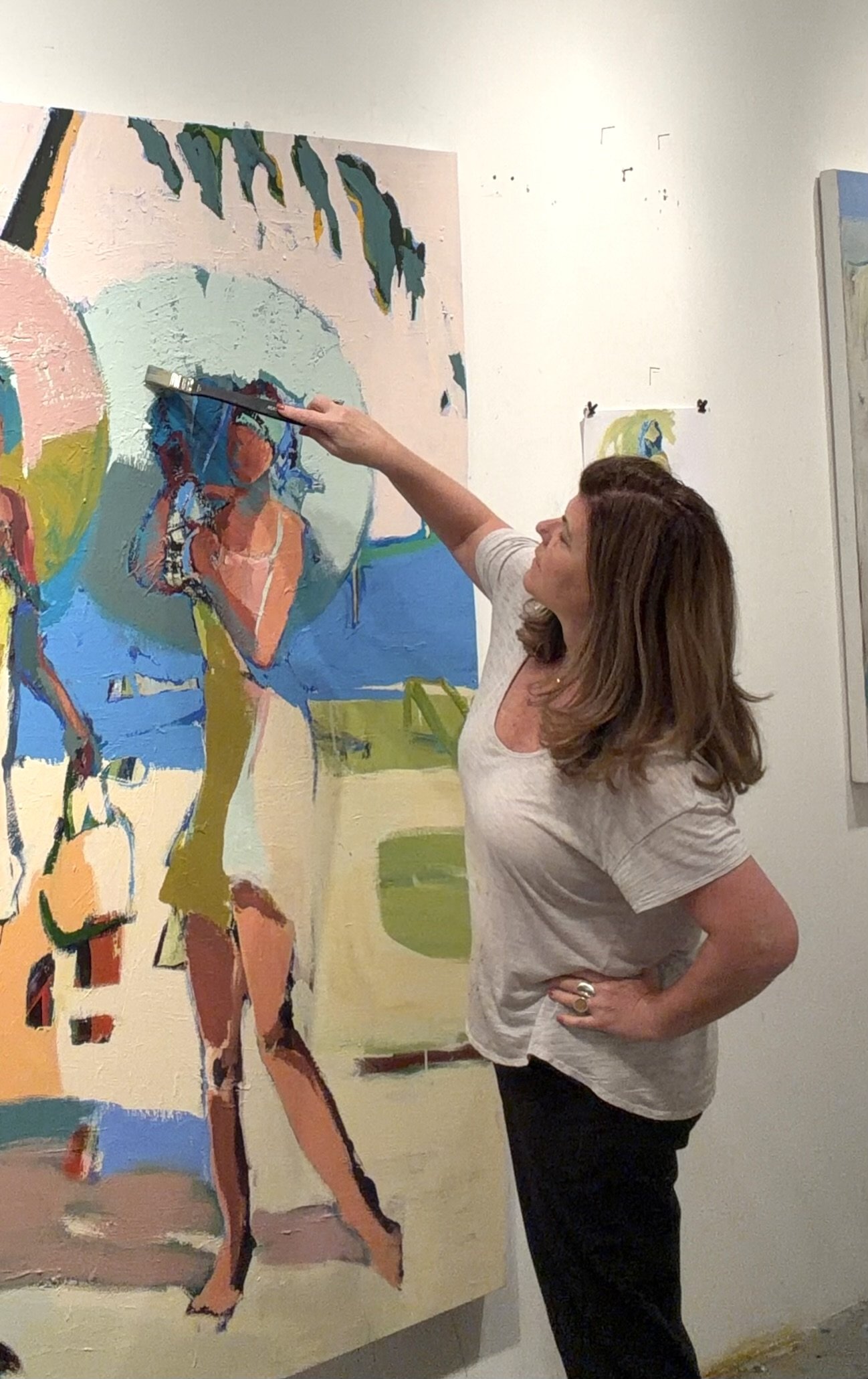 A woman with brown hair, wearing a light gray T-shirt and black pants, standing in front of a colorful painting of a woman in a bikini, shaping or touching the artwork with a large paintbrush in her right hand.