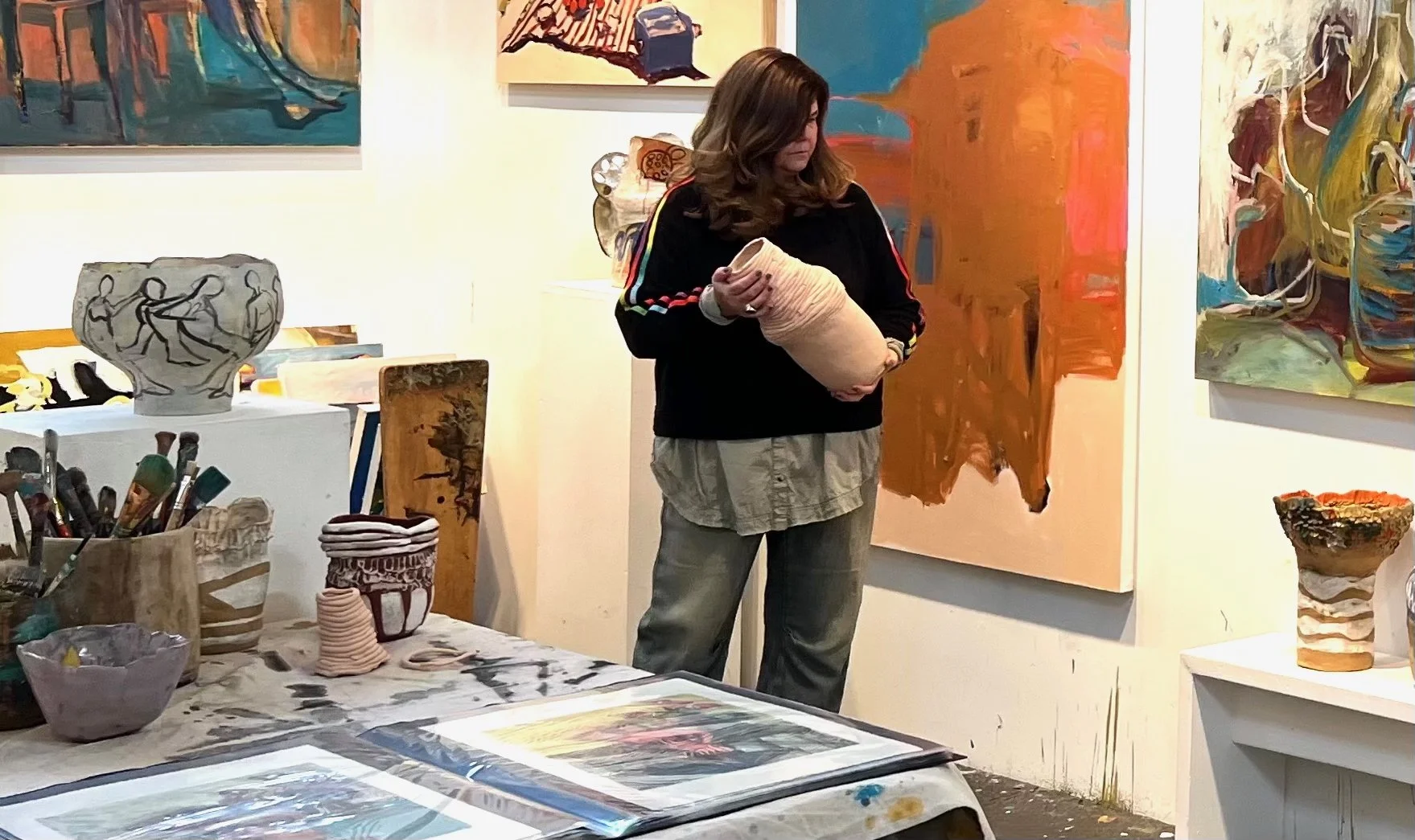 A woman in a dark sweatshirt and beige pants holding a ceramic vessel in an art gallery surrounded by colorful paintings and pottery, with art supplies on a table in the foreground.