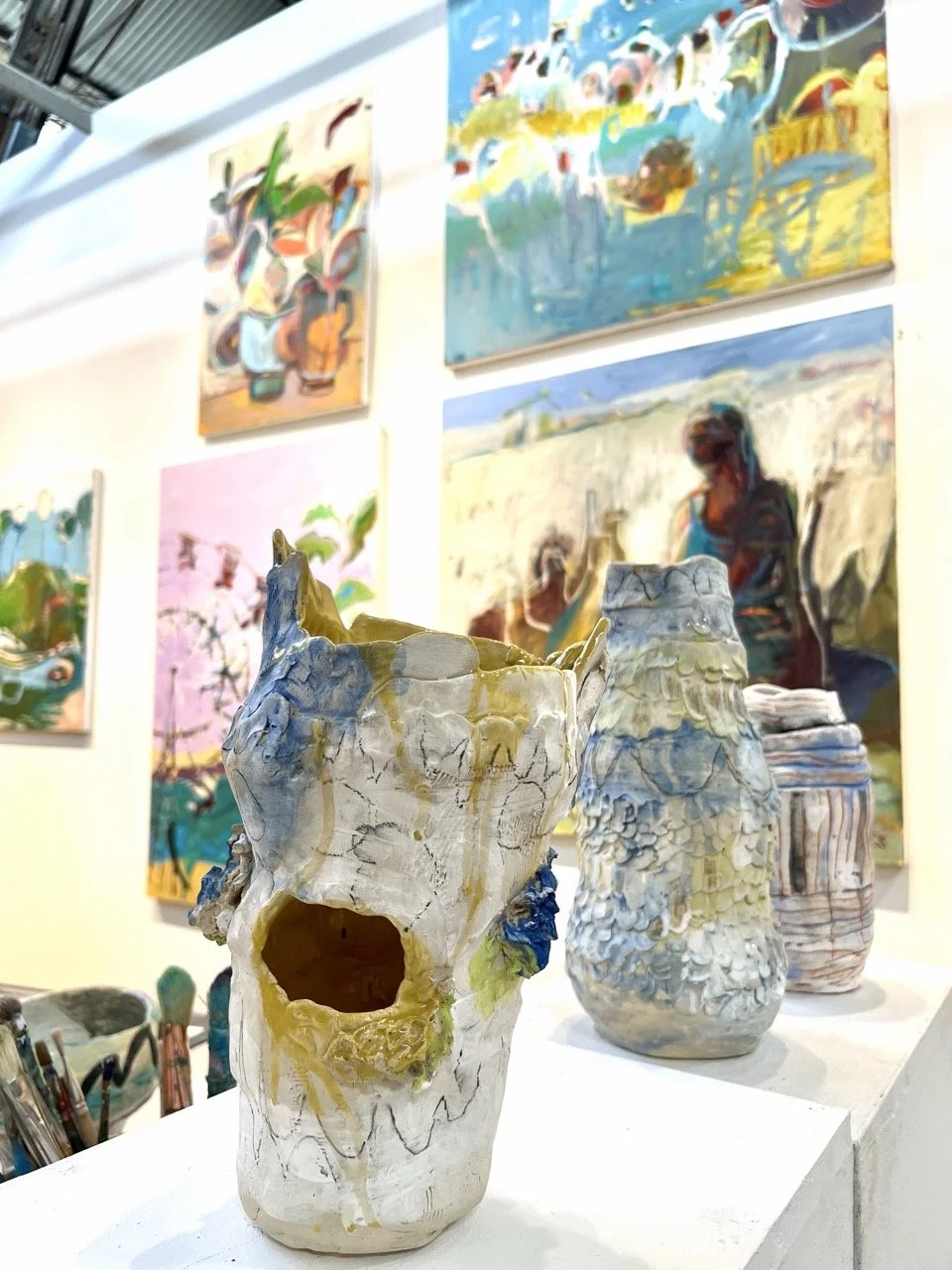 Ceramic sculptures displayed on a white surface at an art gallery, with colorful abstract paintings hanging on the wall behind them.