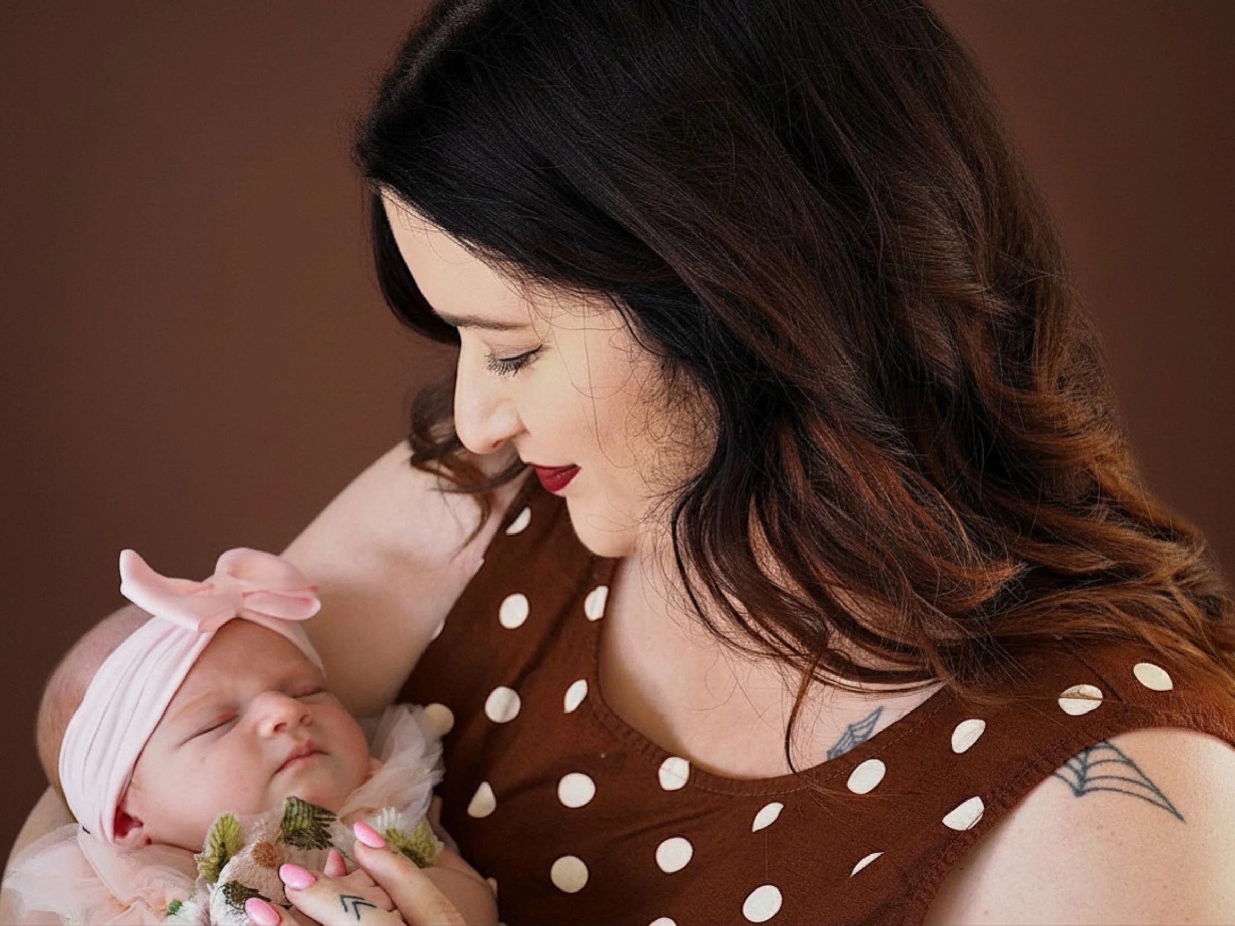 A woman with long dark hair and tattoos gently holding a newborn baby, both lying down against a brown background.