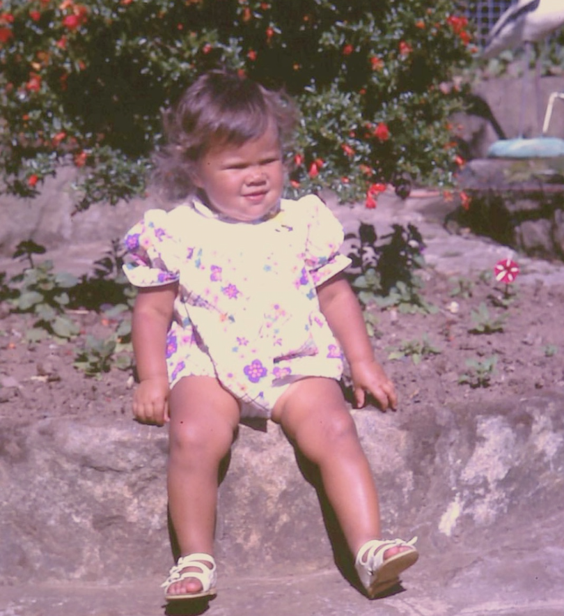 A young girl with short brown hair sitting on a stone ledge outdoors with a flower garden behind her. She is wearing a white dress with colorful floral patterns and yellow sandals, and has a perplexed expression.