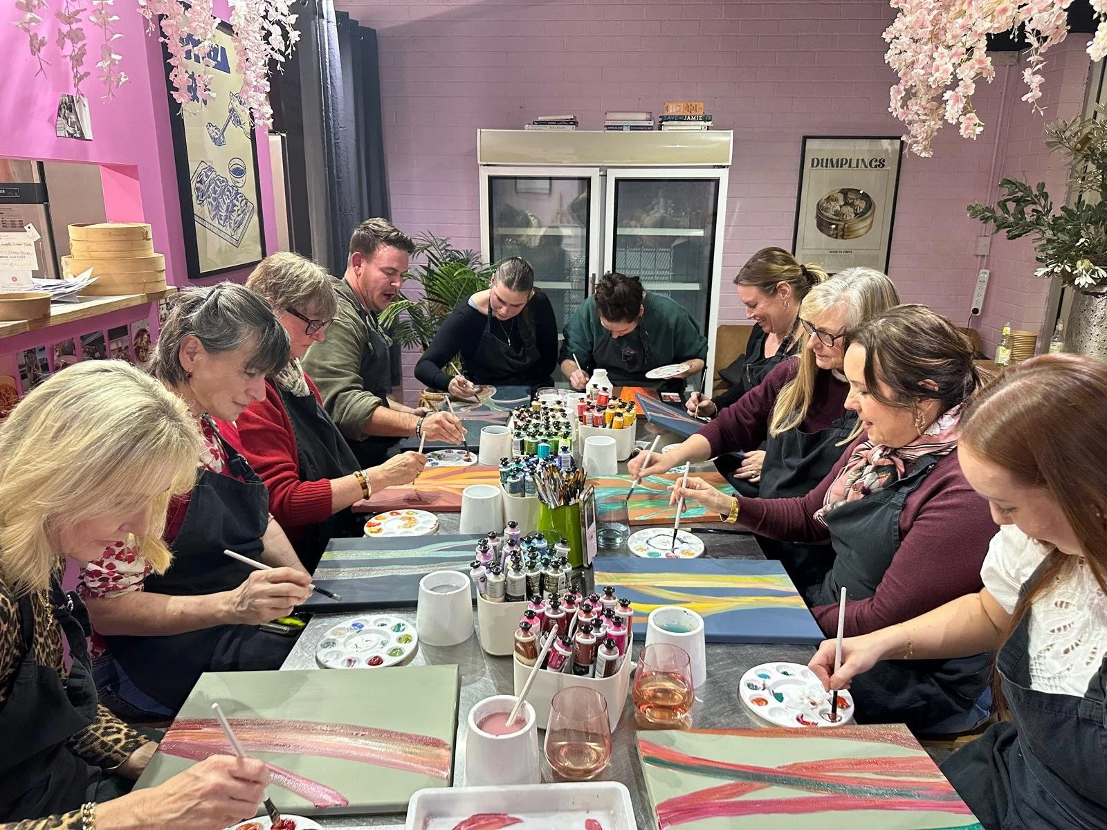 Group of women and one man participating in a painting class, working on colorful abstract paintings at a long table with paints, brushes, and wine glasses in a decorated art studio.