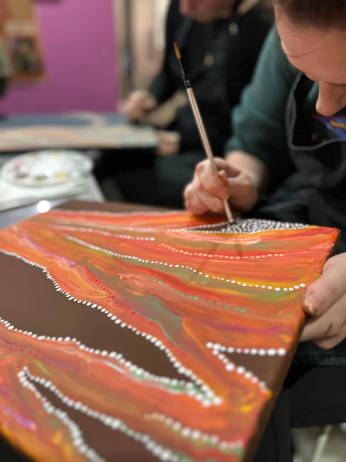 Person painting an abstract artwork with vibrant orange, red, green, purple, and brown colors, outlined with white dots, using a thin brush.