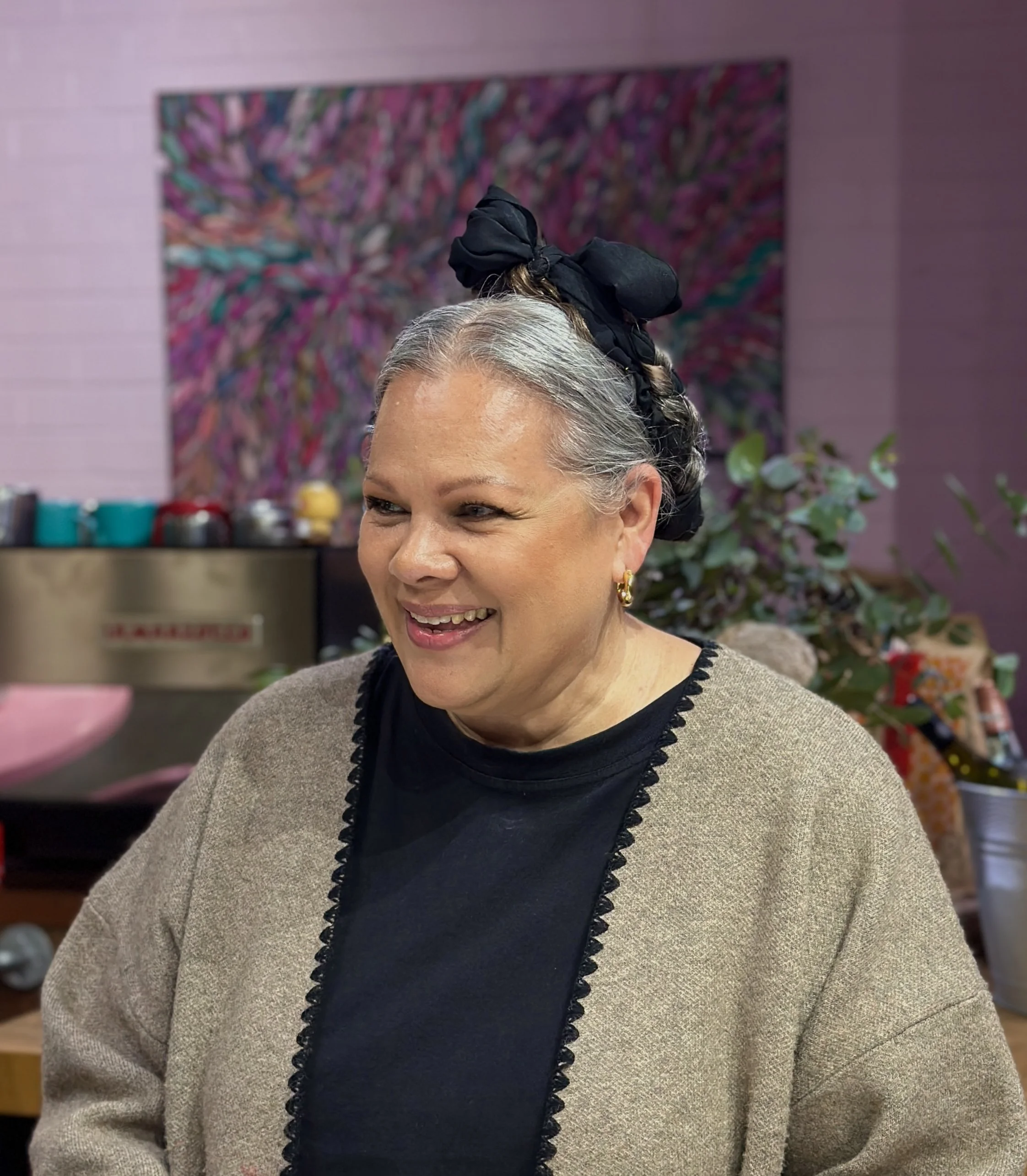 Close-up of a woman with gray hair styled in a braid, wearing a black headscarf and gold earrings, smiling indoors with a colorful abstract painting and plants in the background.