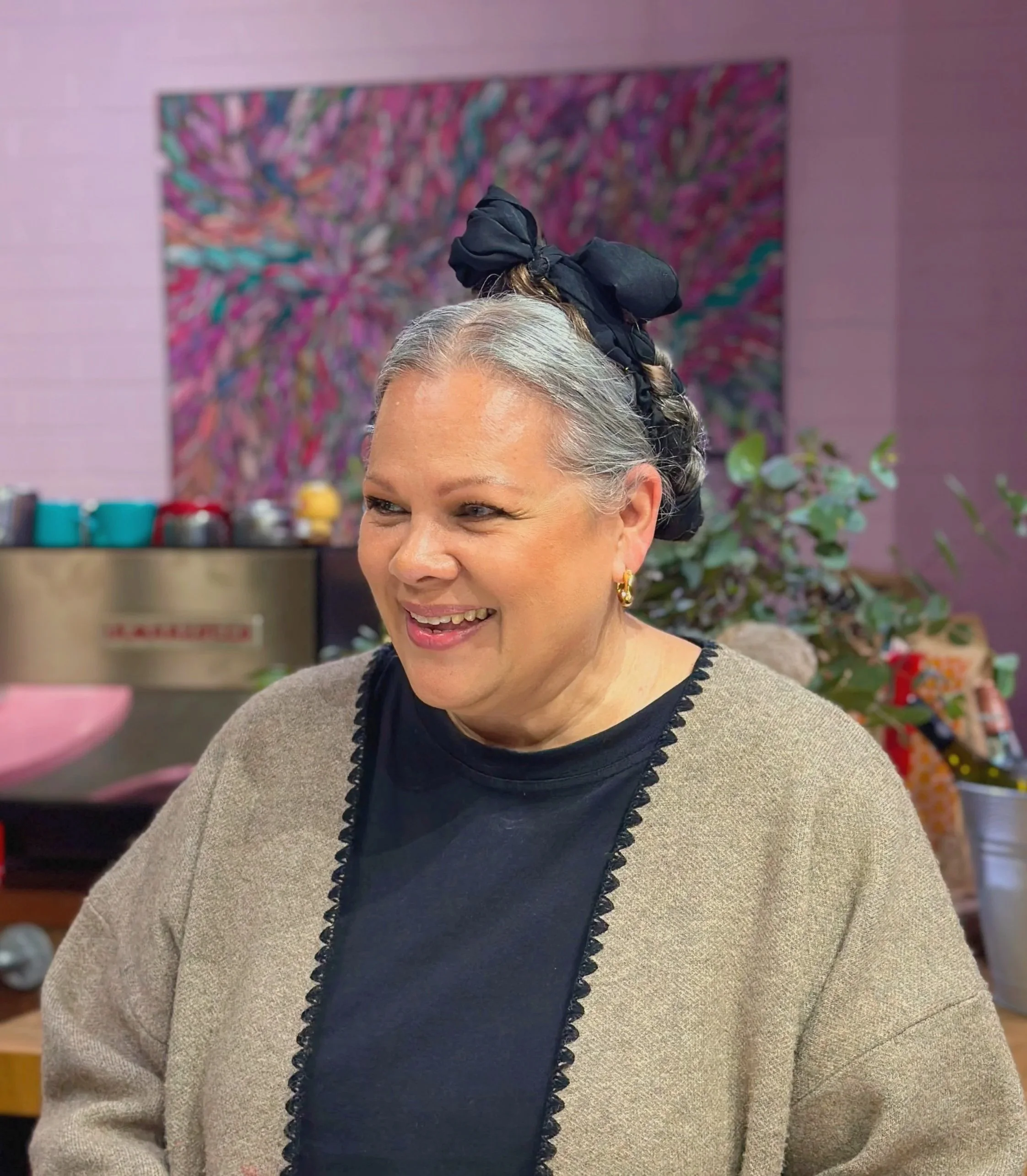 Close-up of a woman with gray hair tied with a black headscarf, wearing a beige cardigan and black top with lace trim, smiling in an indoor setting with colorful artwork and plants in the background.