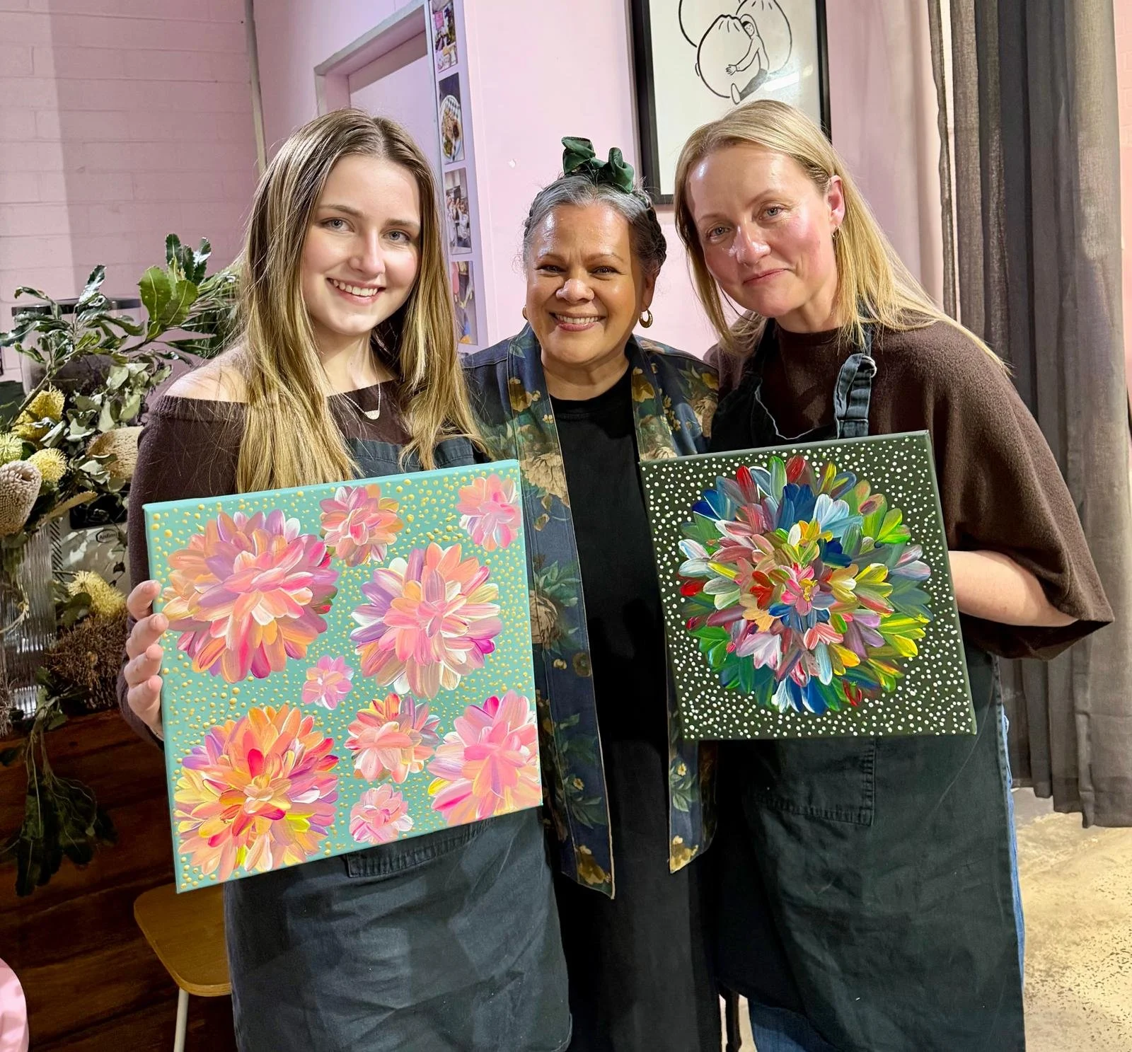 Three women standing indoors, each holding a colorful floral painting, with a pink wall and framed artwork in the background.
