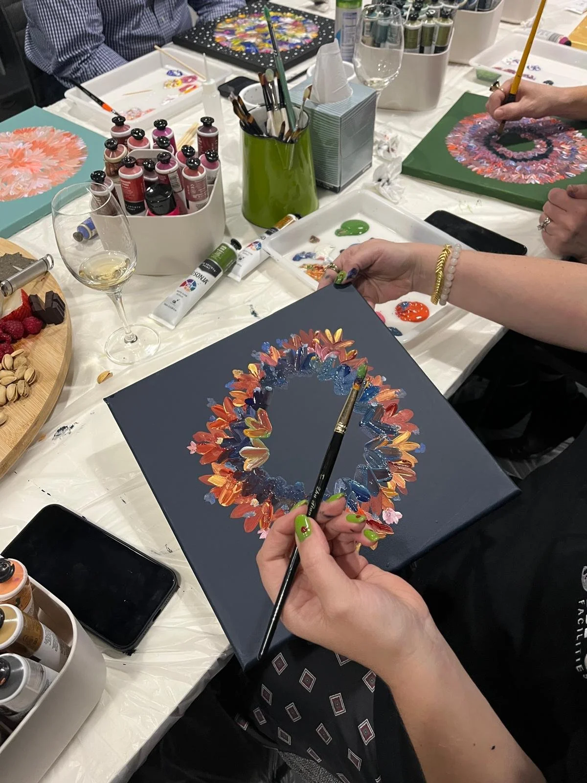 People painting floral wreaths on canvases at a crafting table with paints, brushes, and snacks.