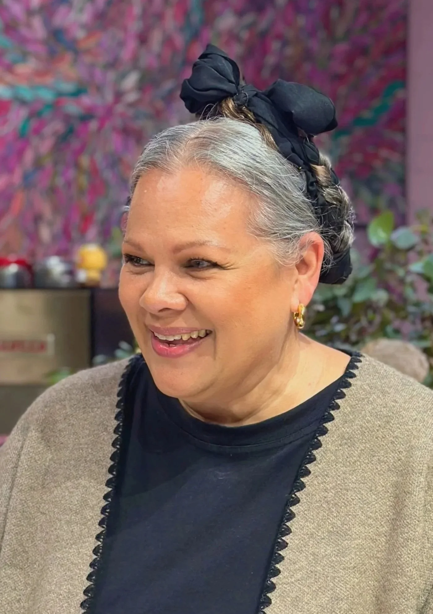 A smiling woman with gray hair tied with a black bow, wearing earrings, a black top, and a beige cardigan, in front of a colorful floral background.