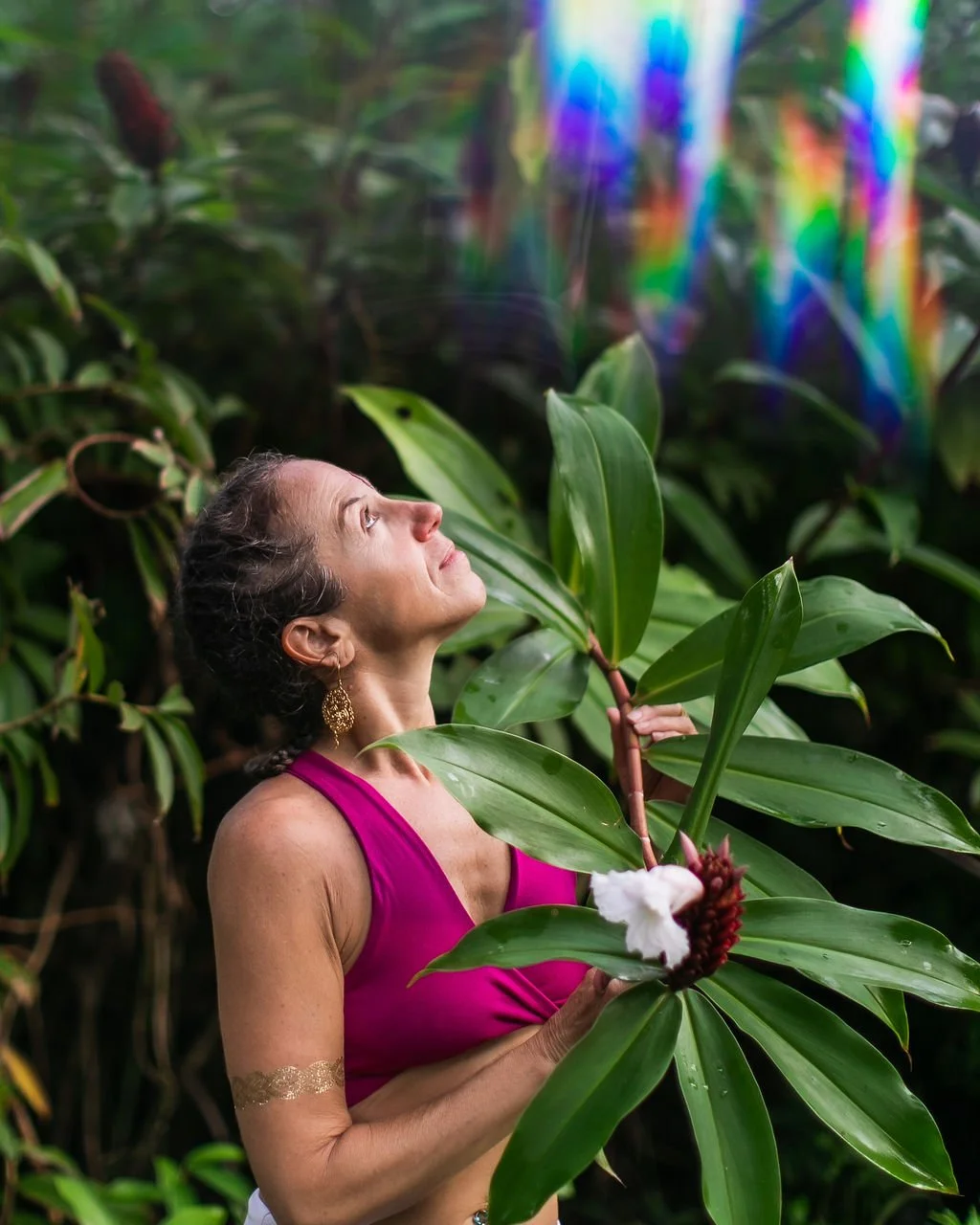 A woman wearing a pink top standing in foliage, looking to the sky.