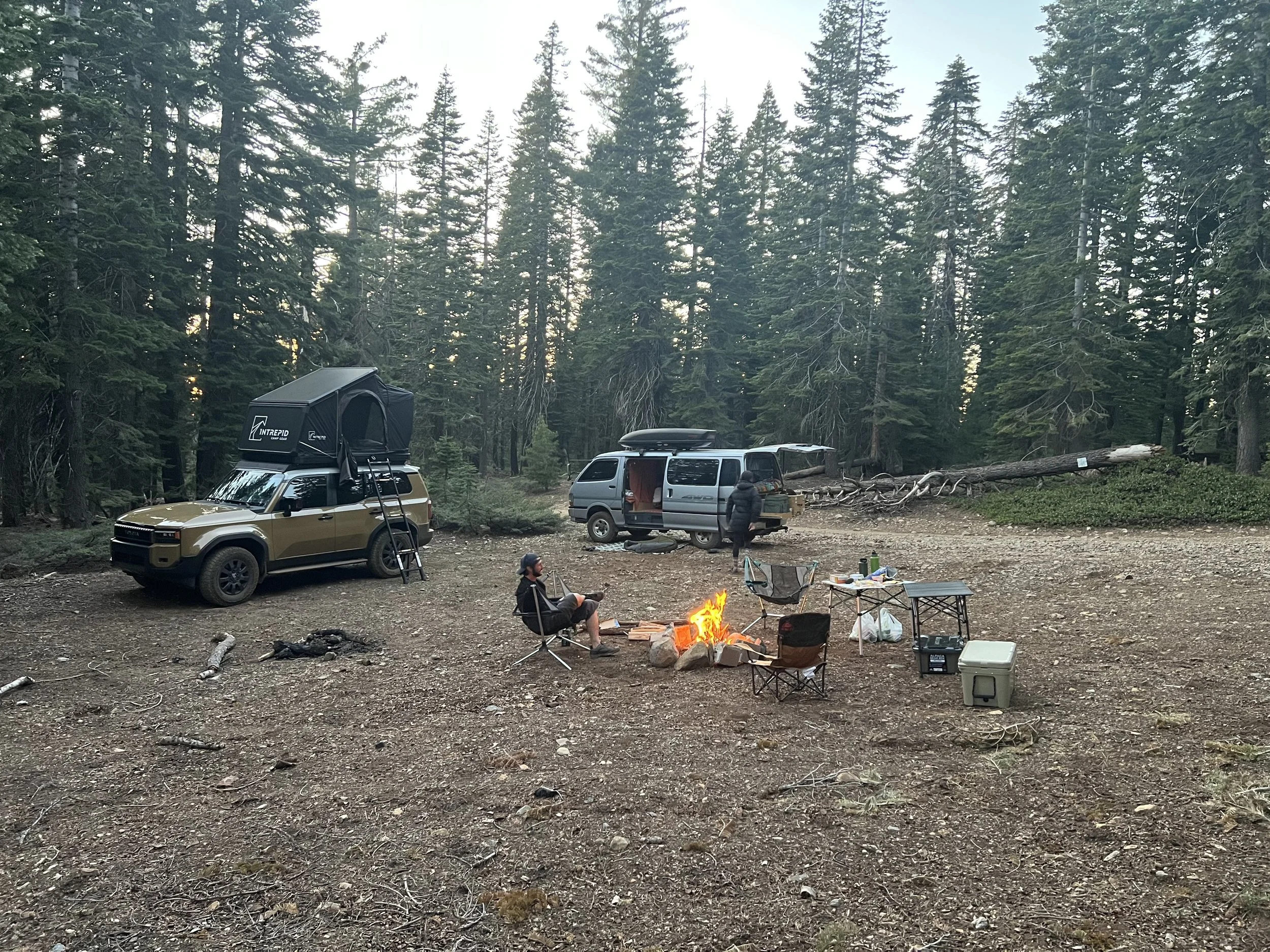 a photo of a man by a fire with vehicles near him on a campground