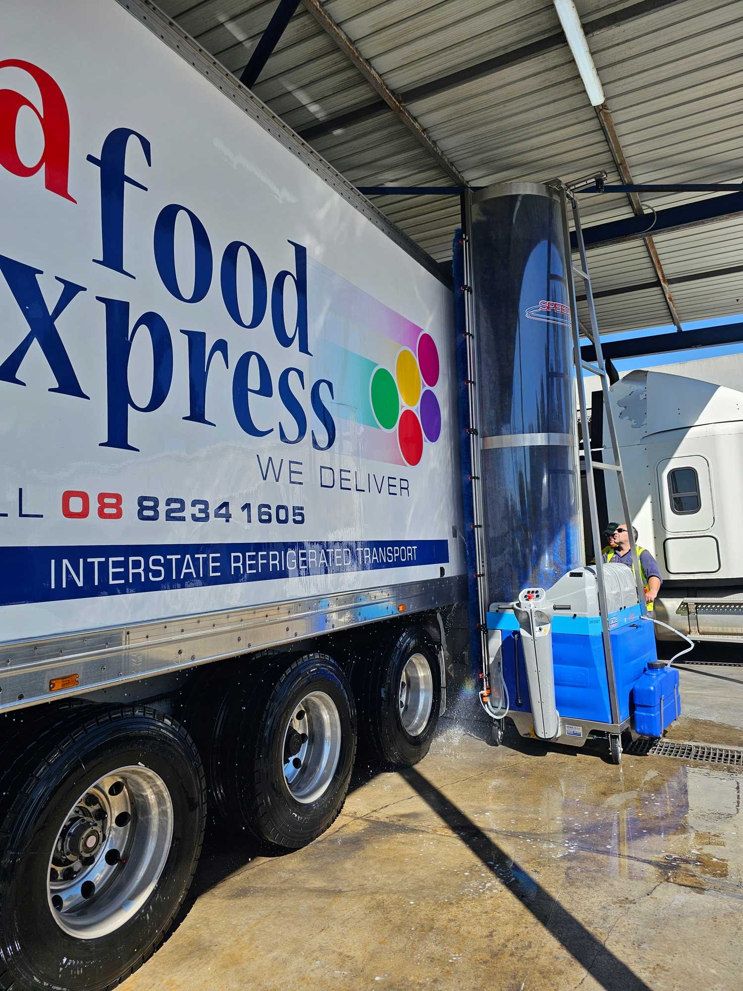 A food supply truck is being cleaned with a high-pressure water hose under a metal roof. The truck is white, with colorful logo and text, and the washing equipment is blue and white.