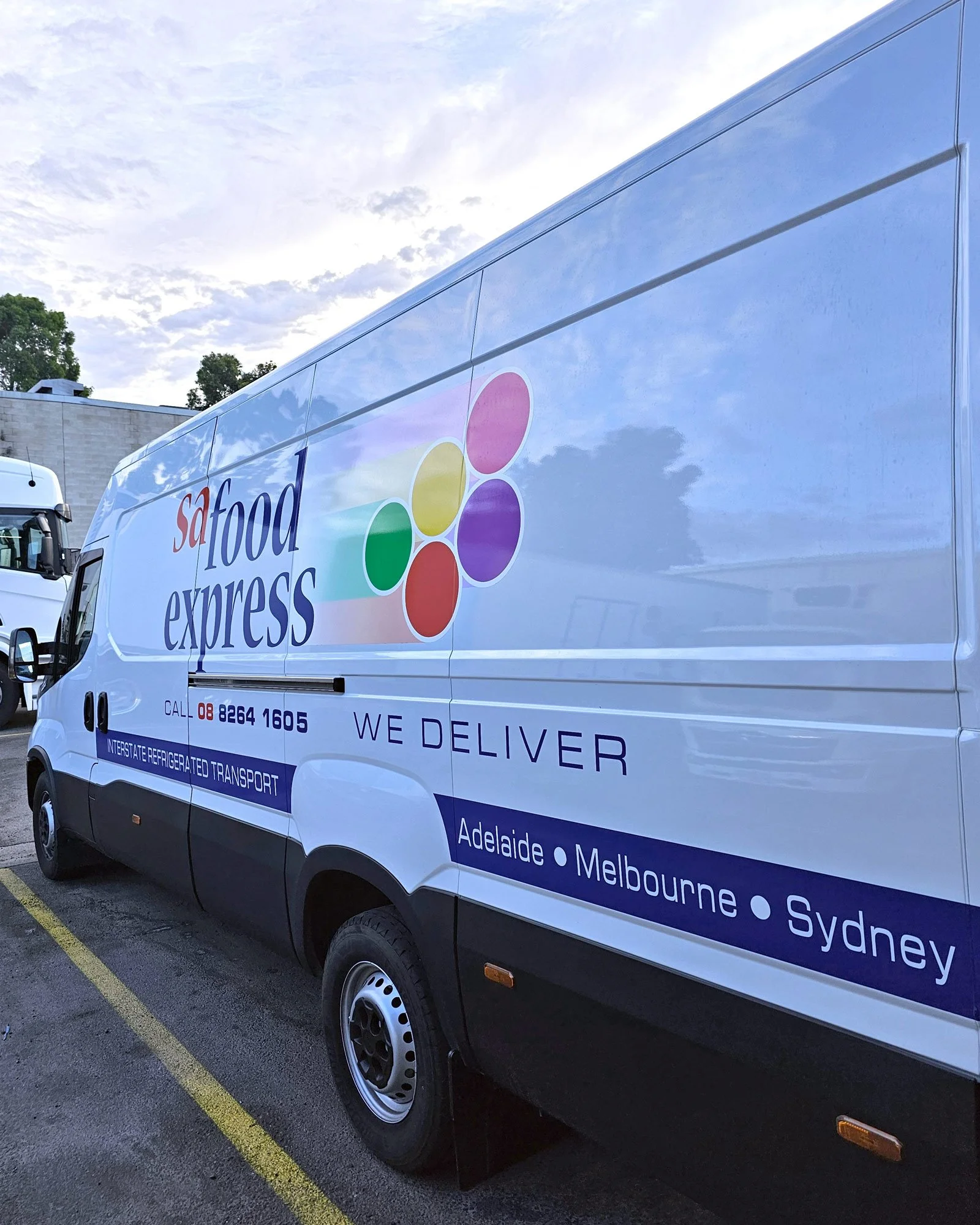 A white delivery truck with colorful logo and text advertising 'Saf Food Express'. The truck is parked in a lot, with a building and trees in the background, under a partly cloudy sky.