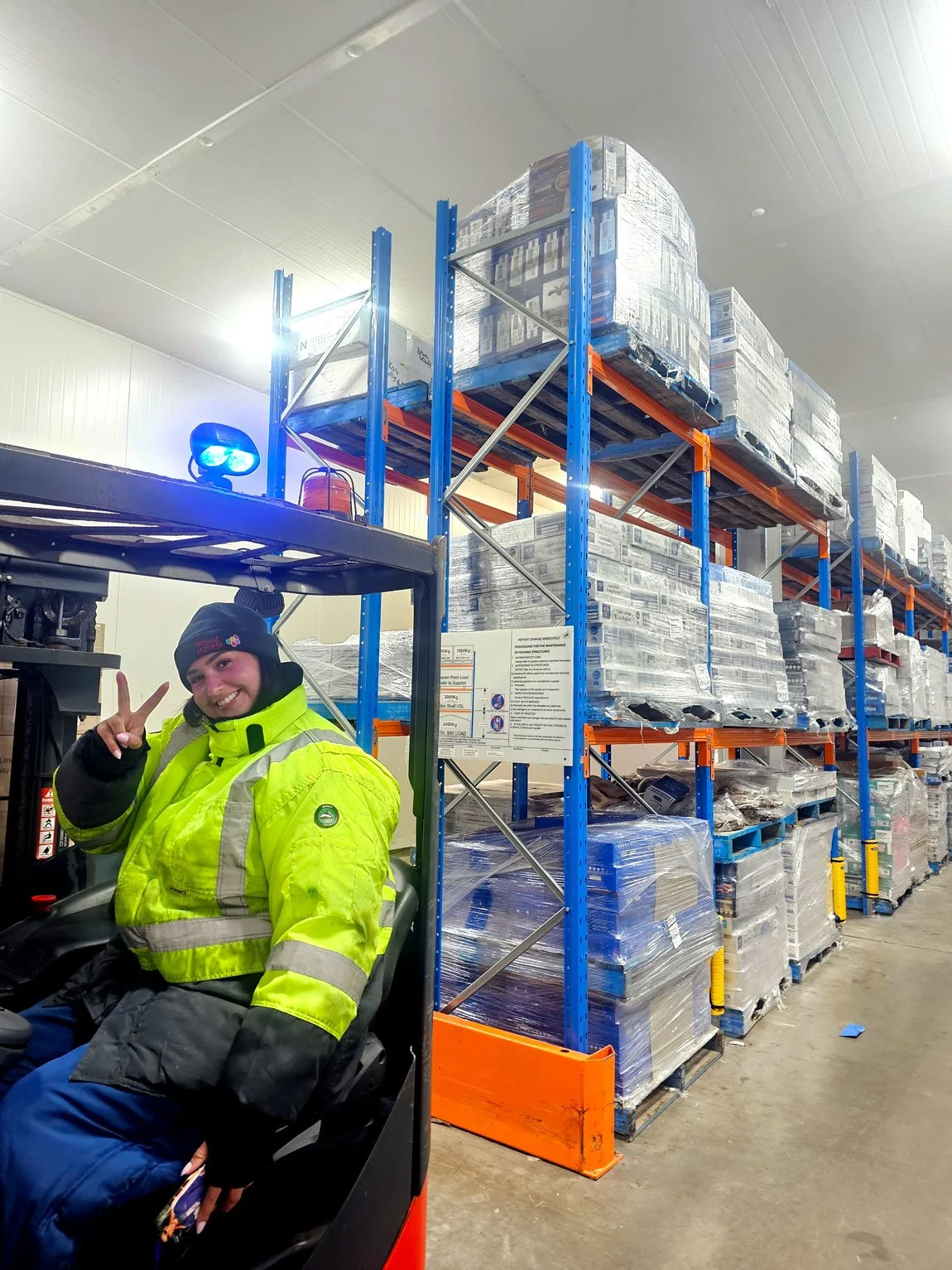 A person with a bright yellow safety jacket and a black beanie is smiling and making a peace sign while sitting in a forklift in a warehouse filled with large stacks of pallets.