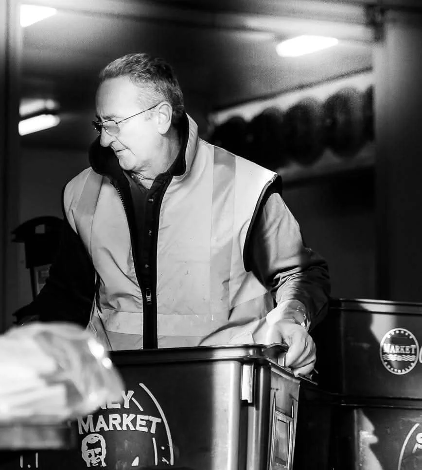 A man wearing glasses and a vest looks down as he pushes a shopping cart at a market.