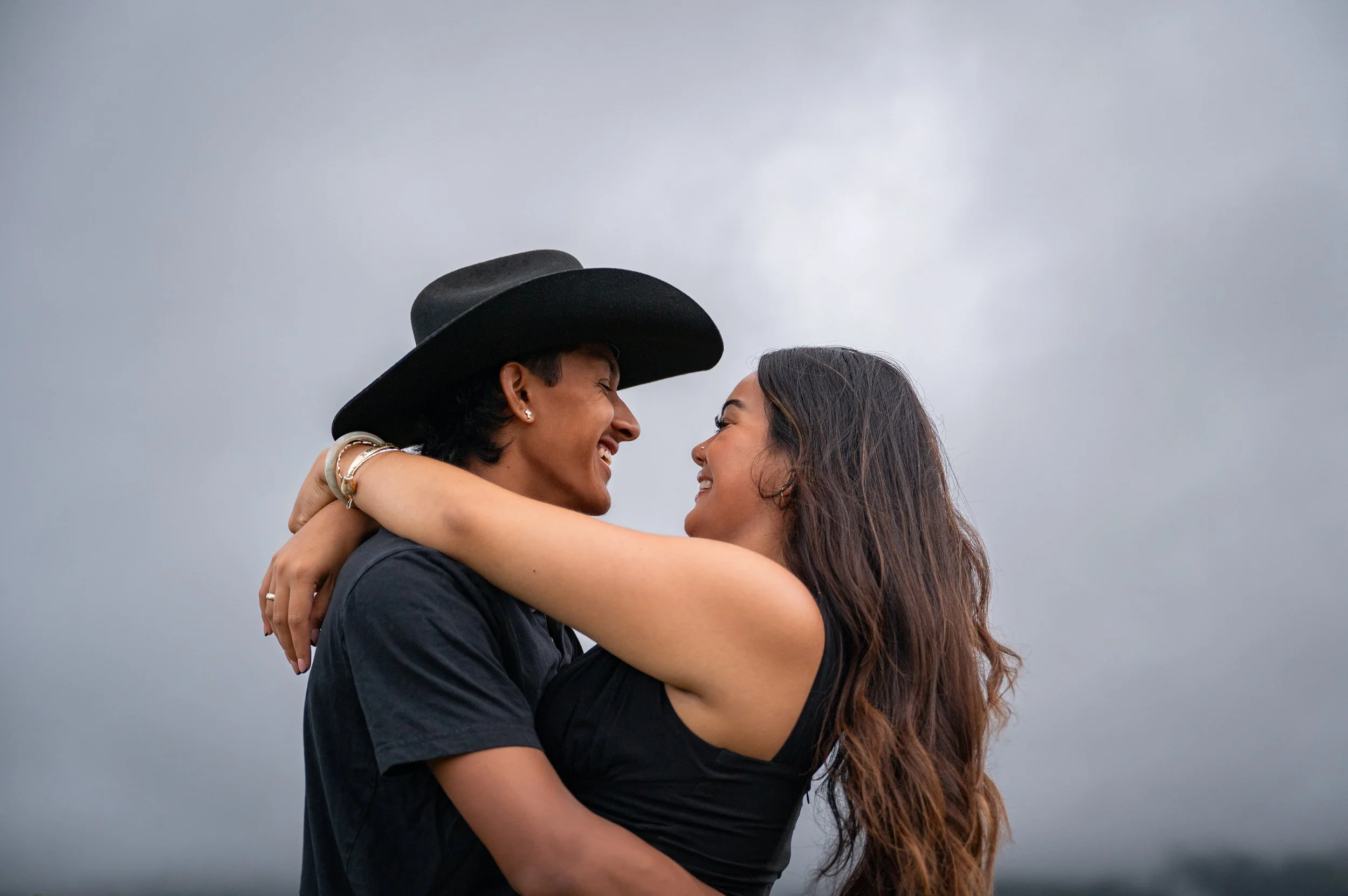 Two women embrace and smile at each other outdoors, with one woman wearing a black wide-brimmed hat and jewelry, and gray cloudy sky in the background.