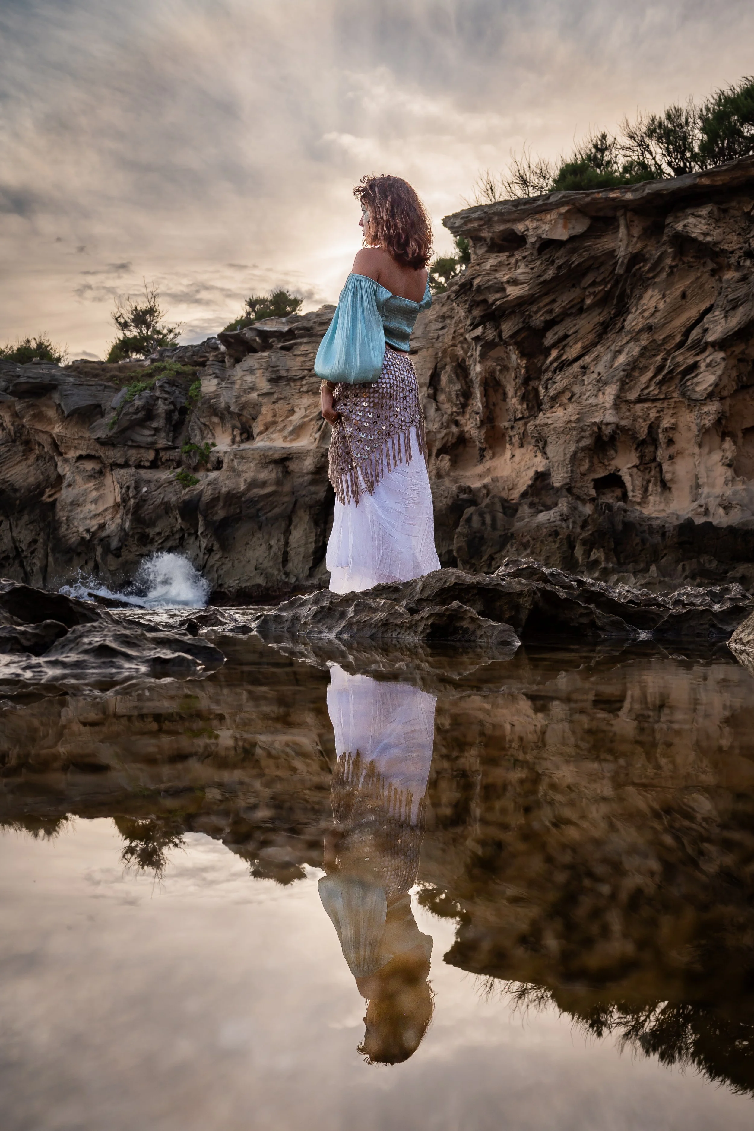A woman in a boho style outfit standing on rocks near the water at sunset or sunrise, with her reflection visible in the water.