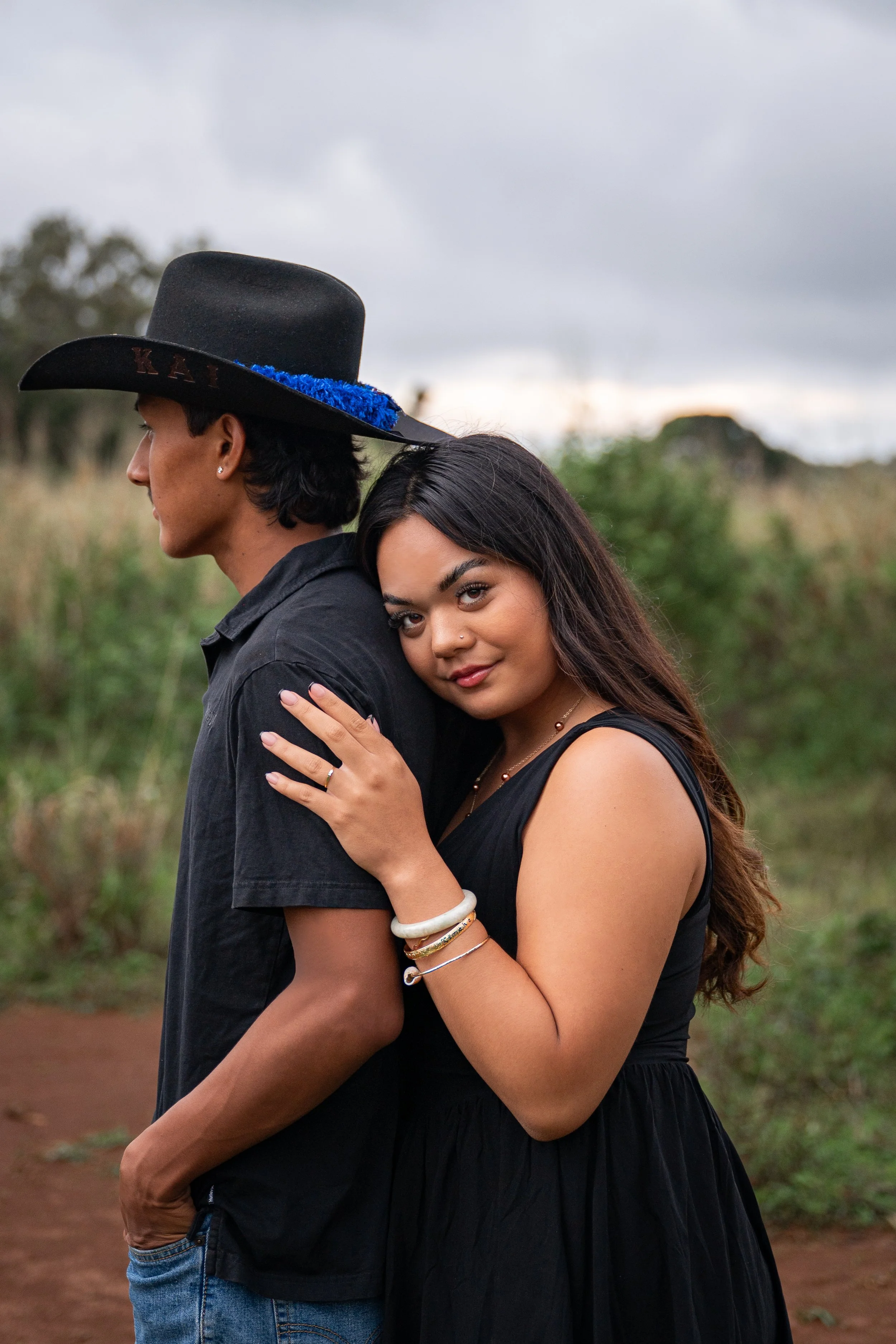 A young man and woman standing outdoors, with the woman hugging the man's back. The man is wearing a black cowboy hat with a blue decoration and a black shirt, and the woman is wearing a black dress and jewelry, smiling at the camera. The background 