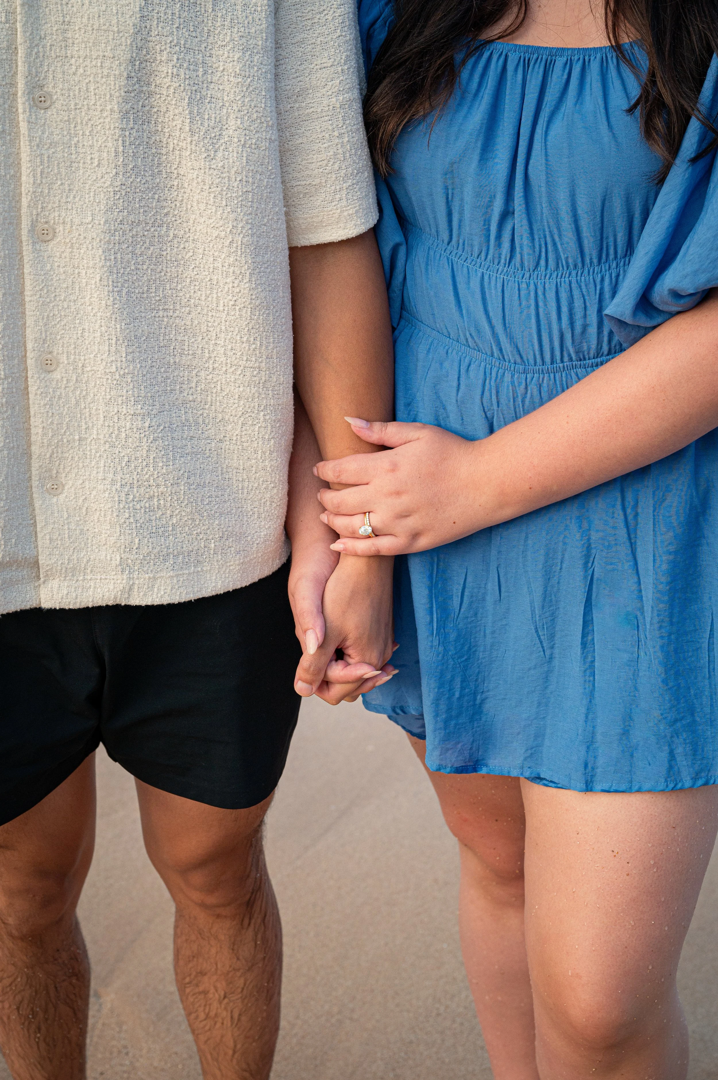 A close-up of two individuals holding hands, one with darker skin and the other with lighter skin, both wearing casual clothing, standing on a sandy surface.