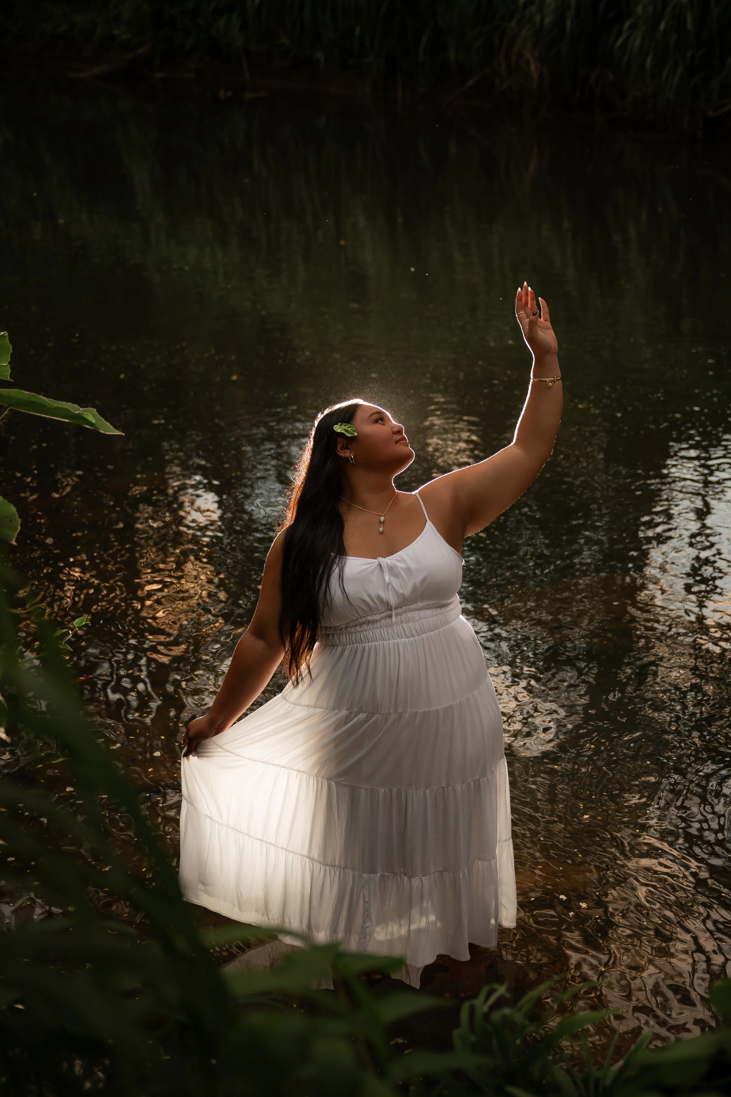 A woman in a white dress standing in a water body, with her right hand raised, surrounded by greenery. The water reflects the sunlight.