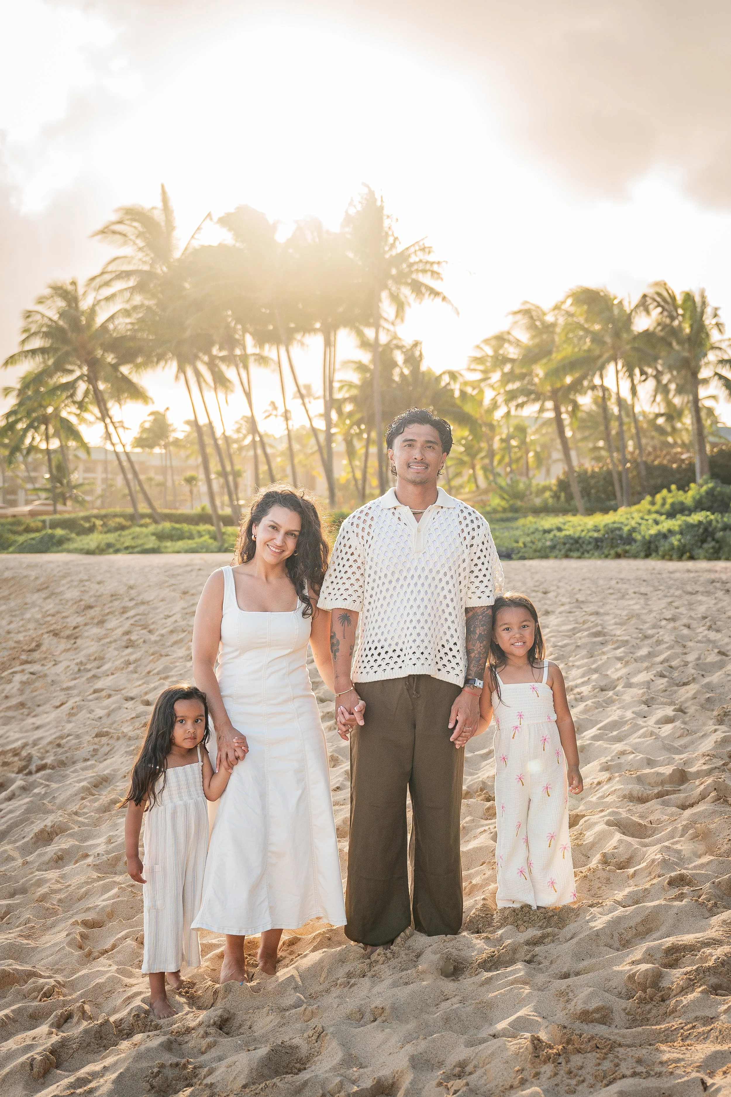 A diverse family of six people standing on a sandy beach with palm trees and a bright sky in the background during sunset.