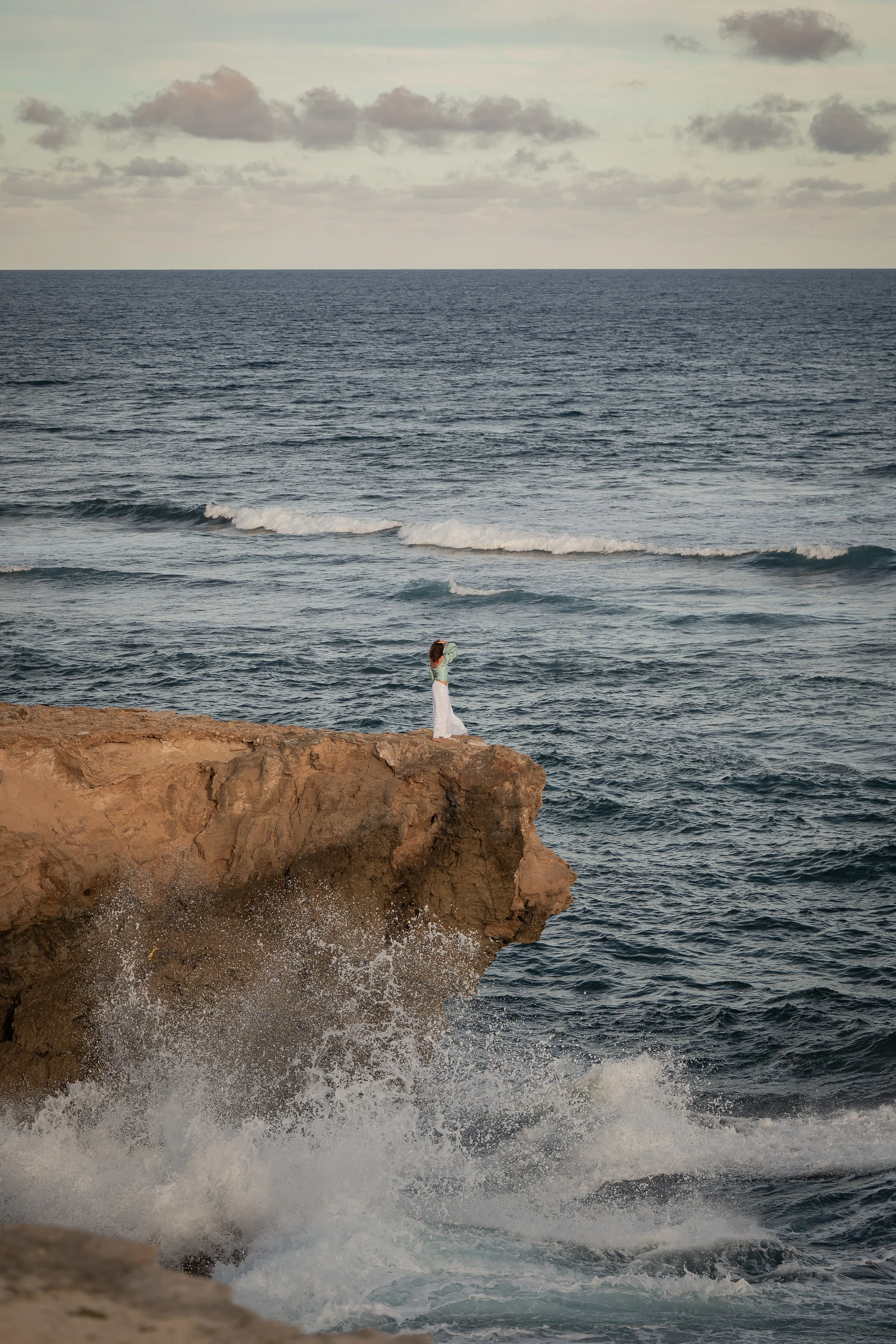 A woman in a white dress standing on a rocky cliff by the ocean, with waves crashing against the rocks. The sky is cloudy with some breaks in the clouds.