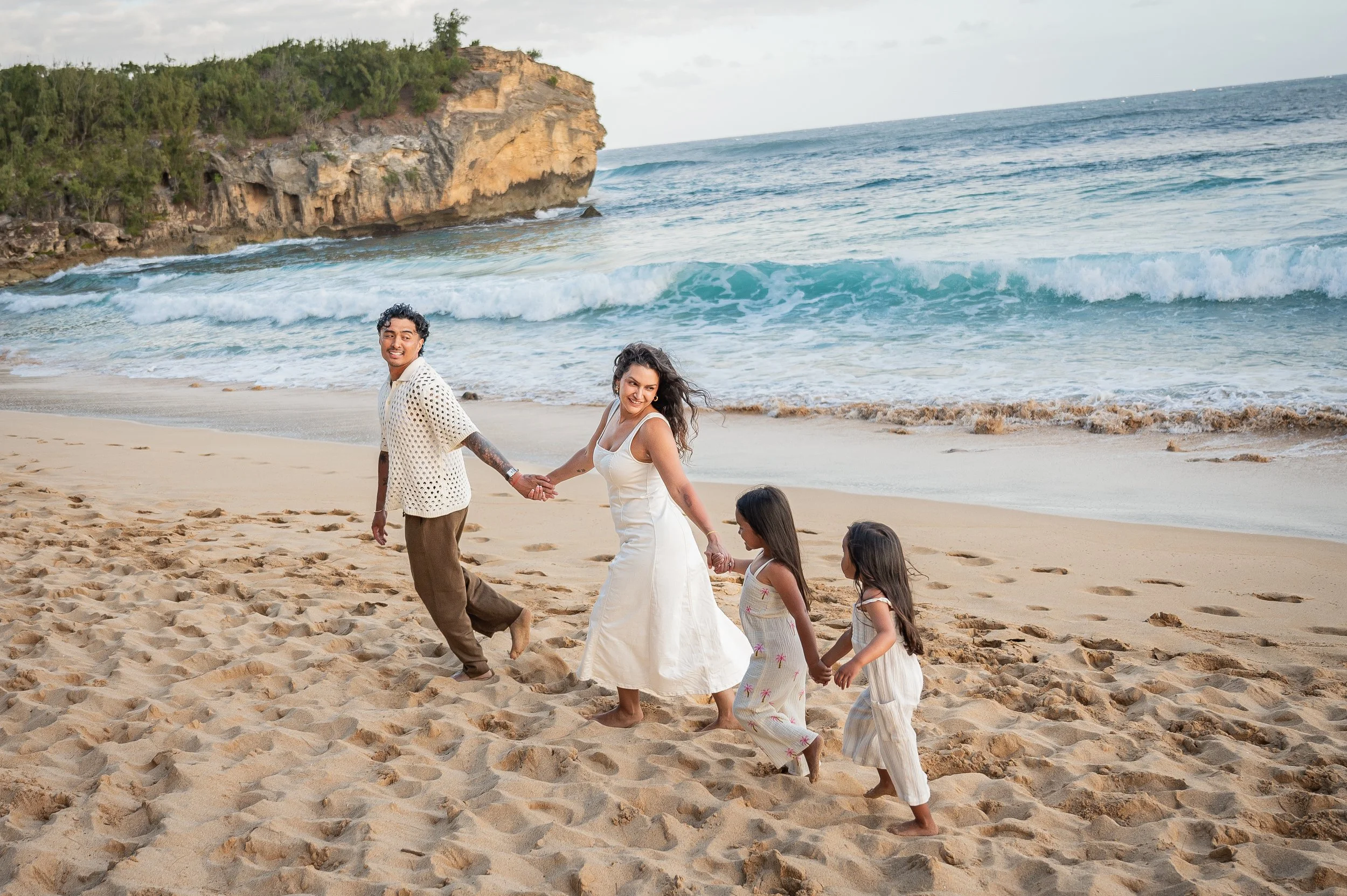 A happy couple holding hands and walking along the beach with three young girls, with ocean waves and a rocky cliff in the background.