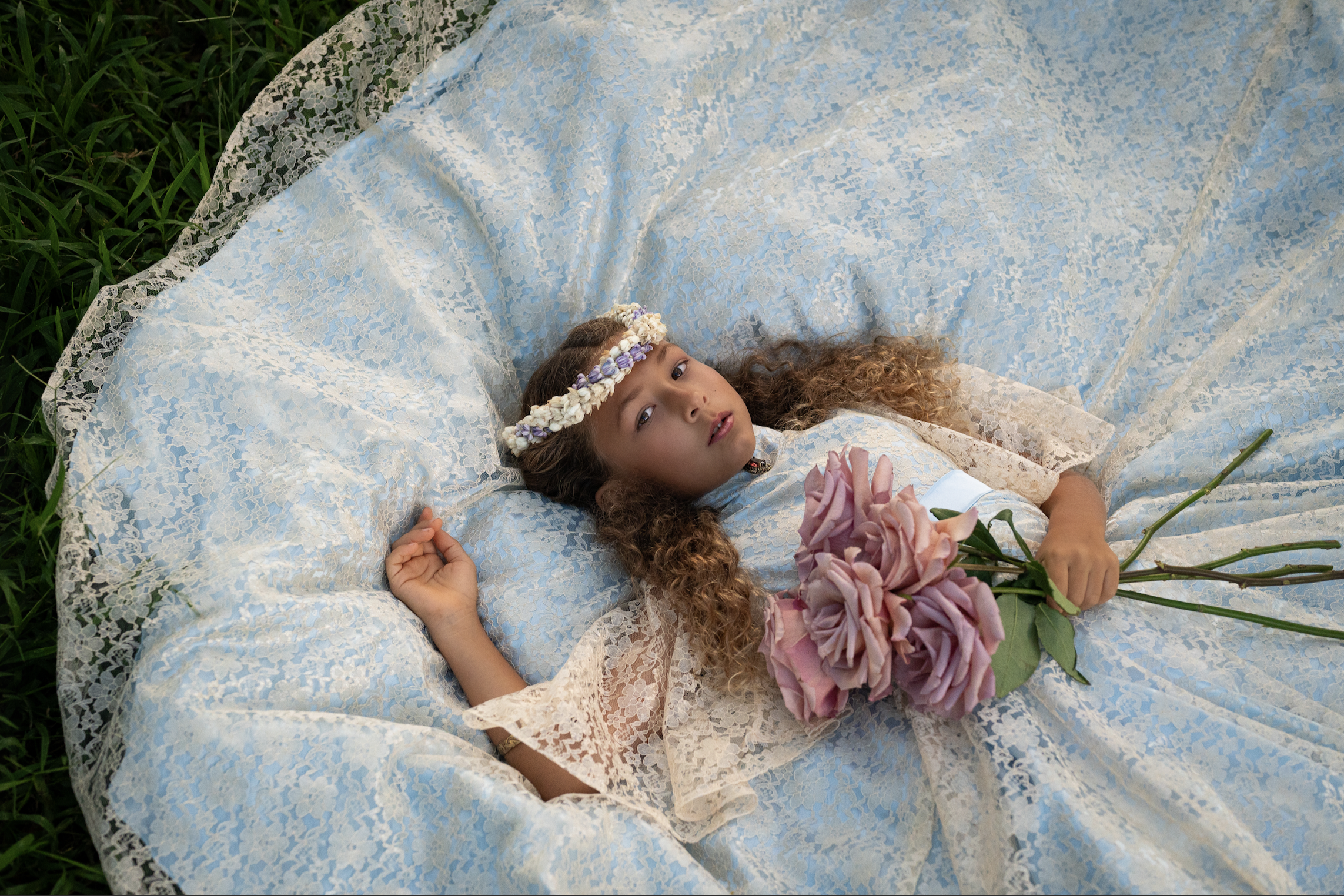 A young girl with curly hair lying on a lace-covered blanket outdoors, wearing a floral crown and holding pink flowers.