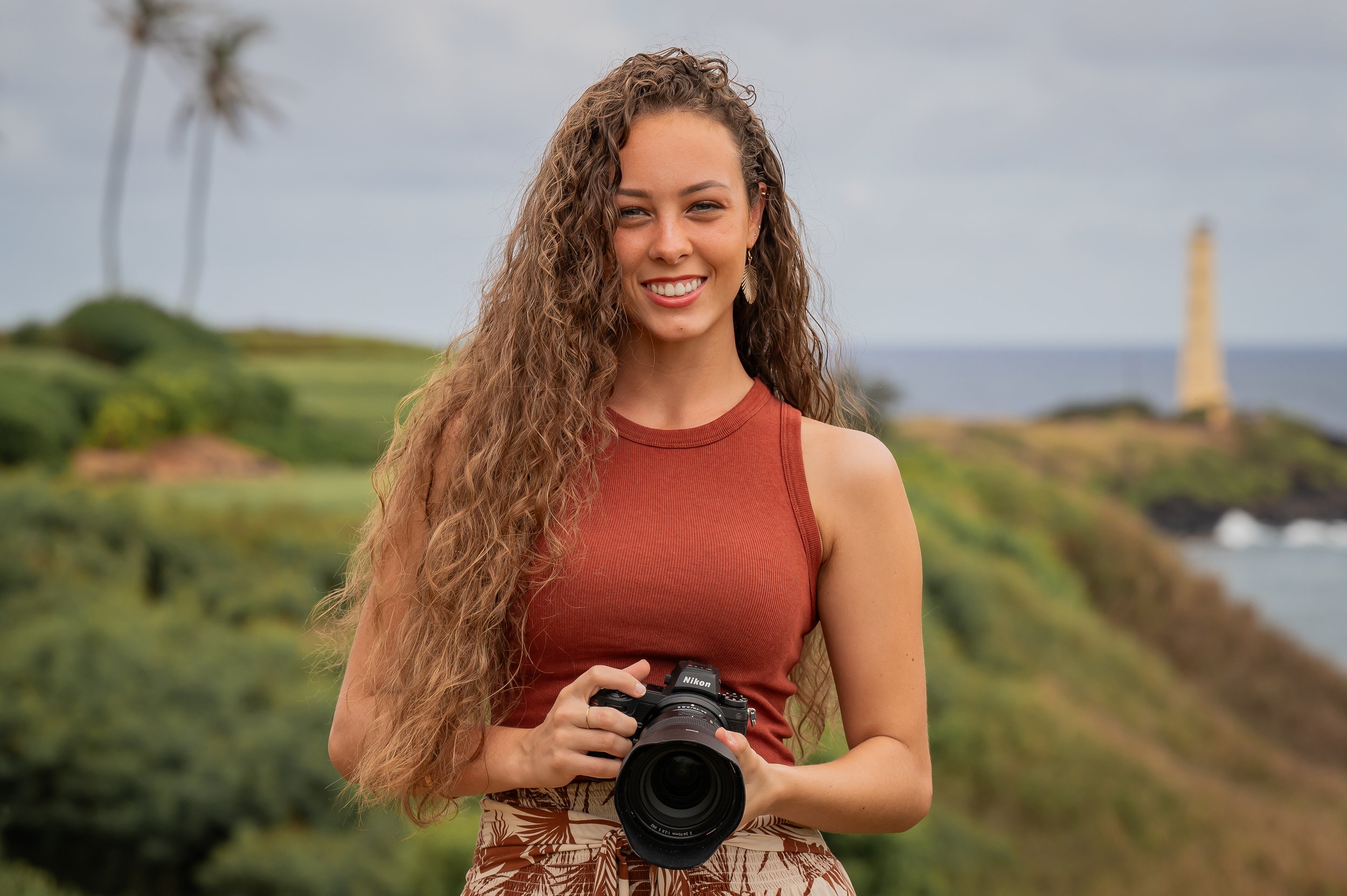 A young woman with long, curly hair smiling and holding a camera outdoors with greenery and a lighthouse in the background.