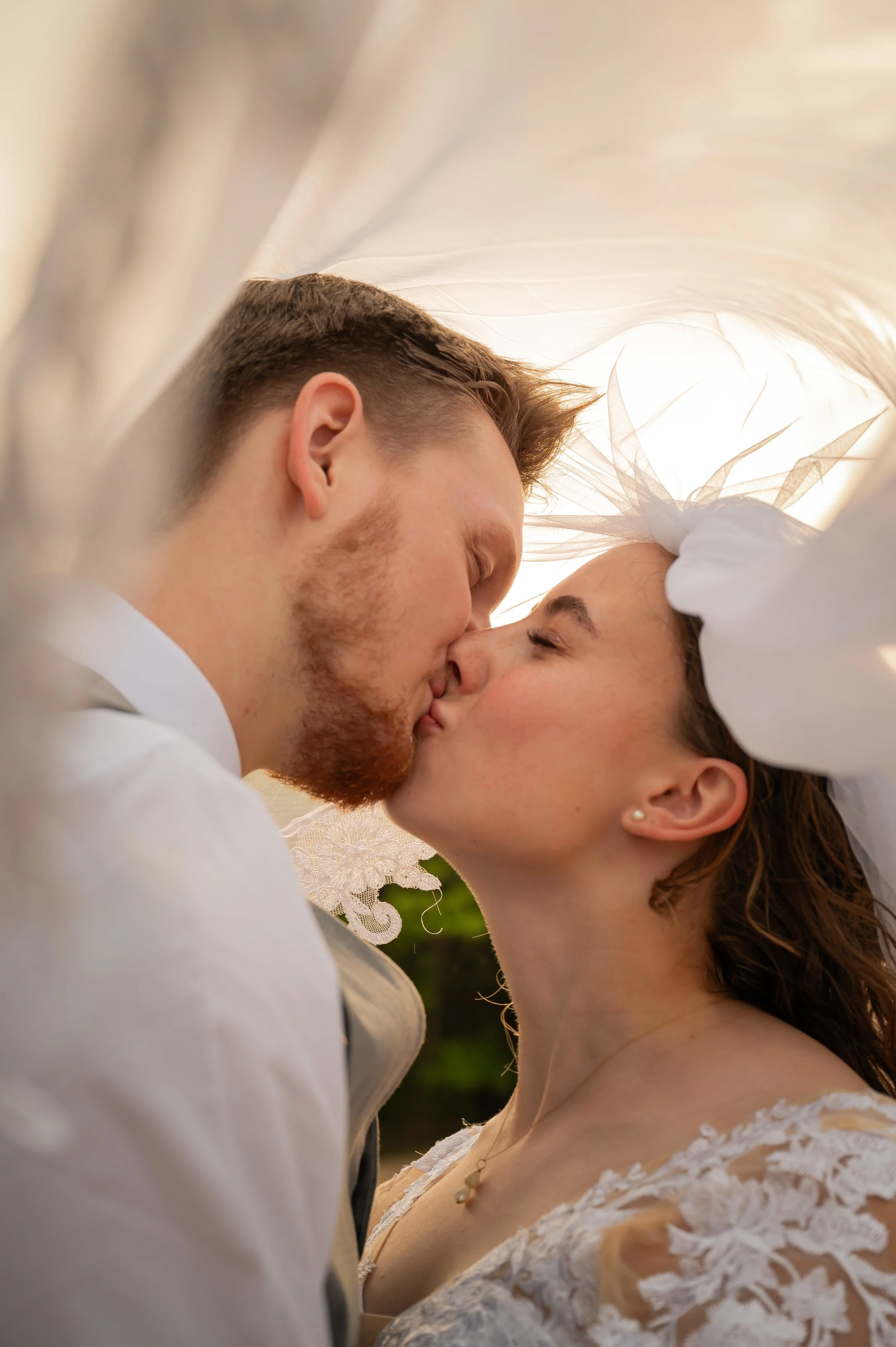 A man and woman are sharing a kiss at their wedding. The man has light brown hair and a beard, and the woman has dark brown hair styled in an updo, wearing earrings, a necklace, and a lace wedding dress. They are surrounded by white fabric, possibly 