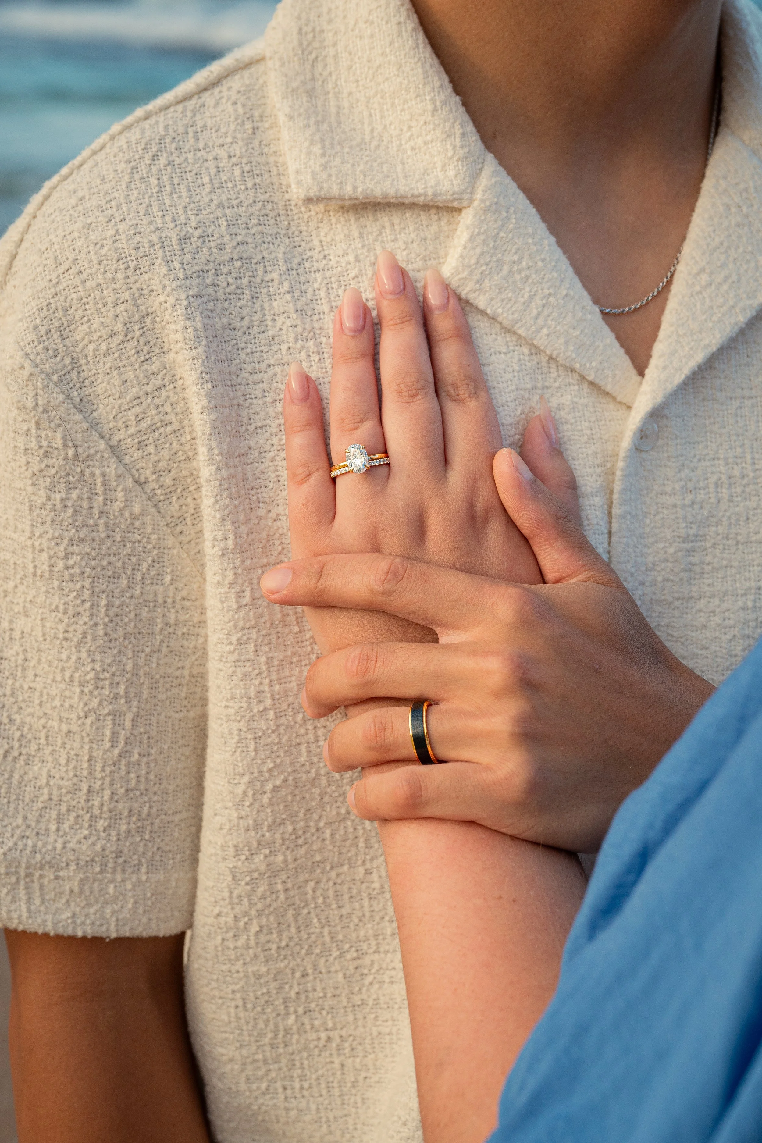 A close-up of two people embracing, with one person's hand showing a diamond engagement ring resting on the other's chest, both wearing casual clothing, near a body of water.