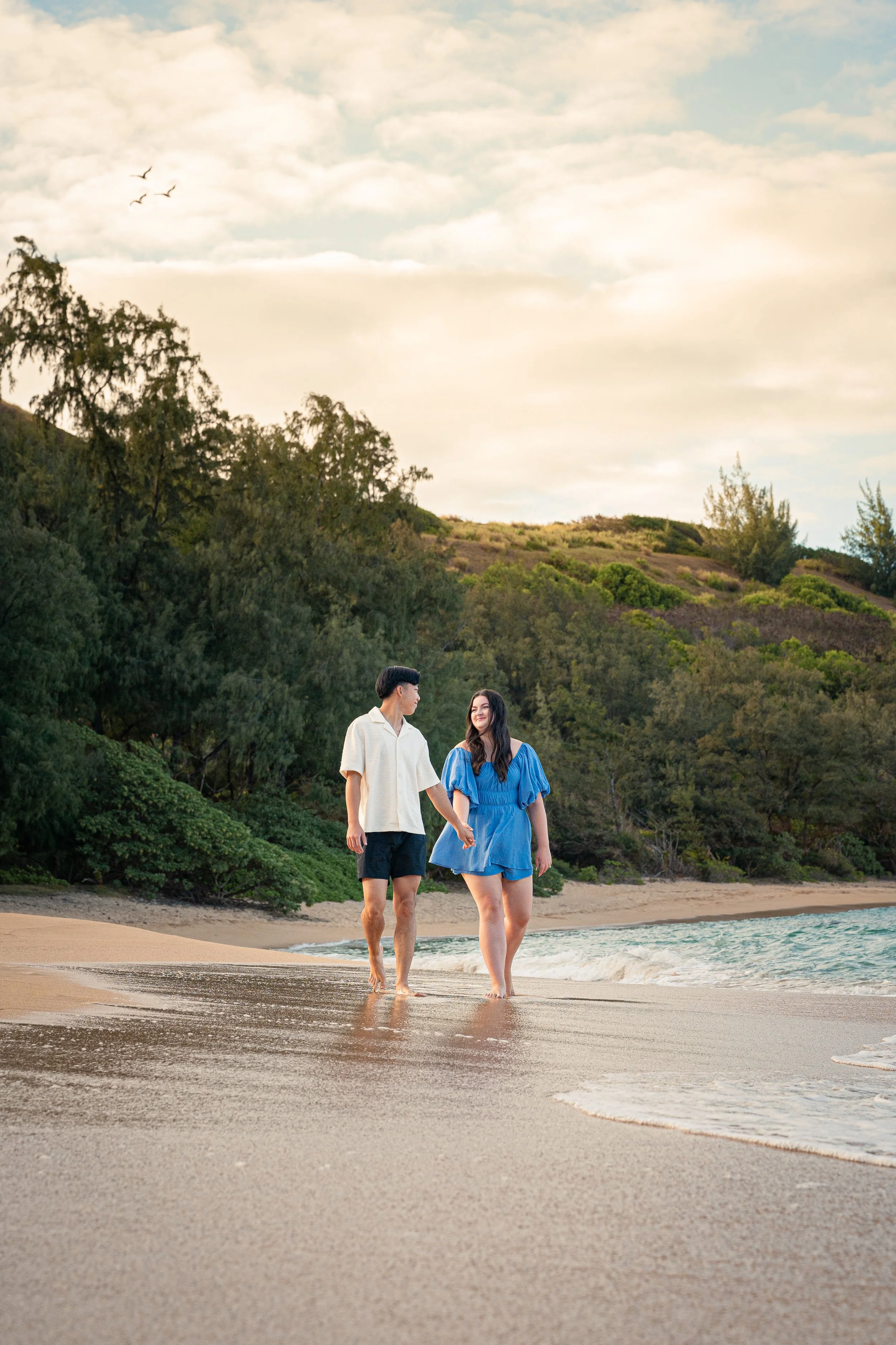 A young couple walking hand in hand along the beach at sunset, with the ocean waves touching their feet and a backdrop of trees and rolling hills.