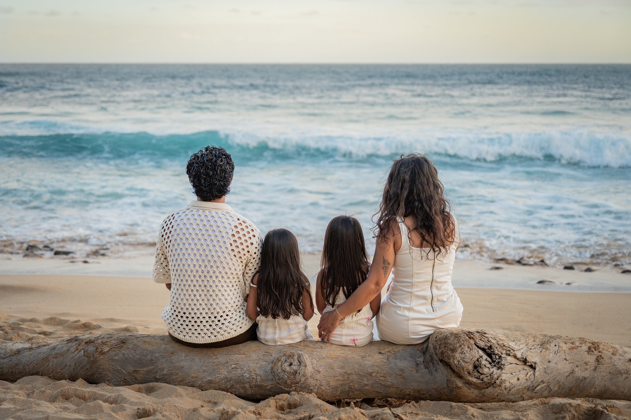 A group of five people, including two adults and three children, sitting on a piece of driftwood on a sandy beach, looking at the ocean waves during sunset or late afternoon.