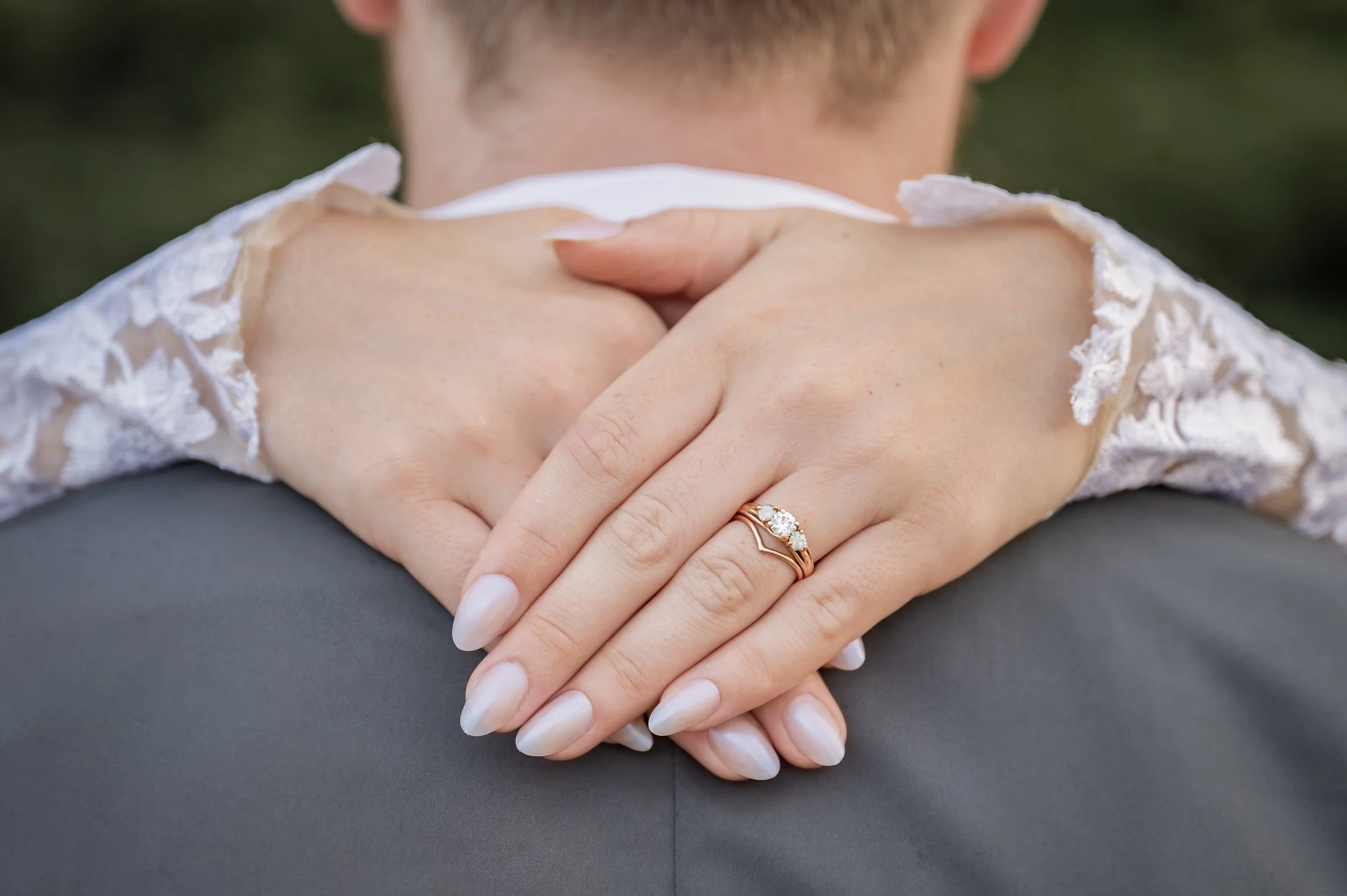 A person with light skin is holding their hands together, wearing a ring with three diamonds on the ring finger of their left hand. They are dressed in a white lace garment, and the background is blurred with some greenery.