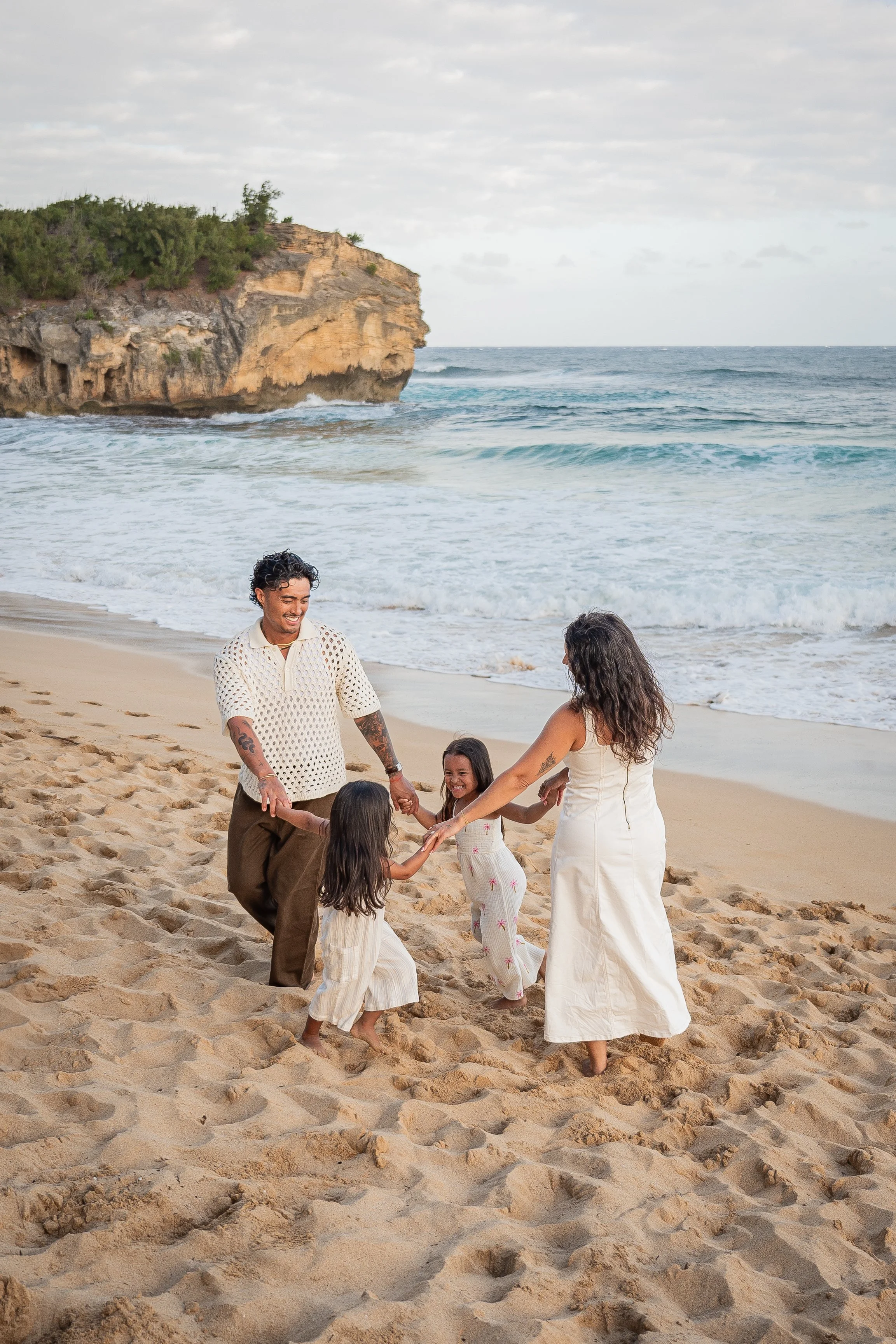 A family dancing and playing on a sandy beach with ocean waves and a rocky cliff in the background.
