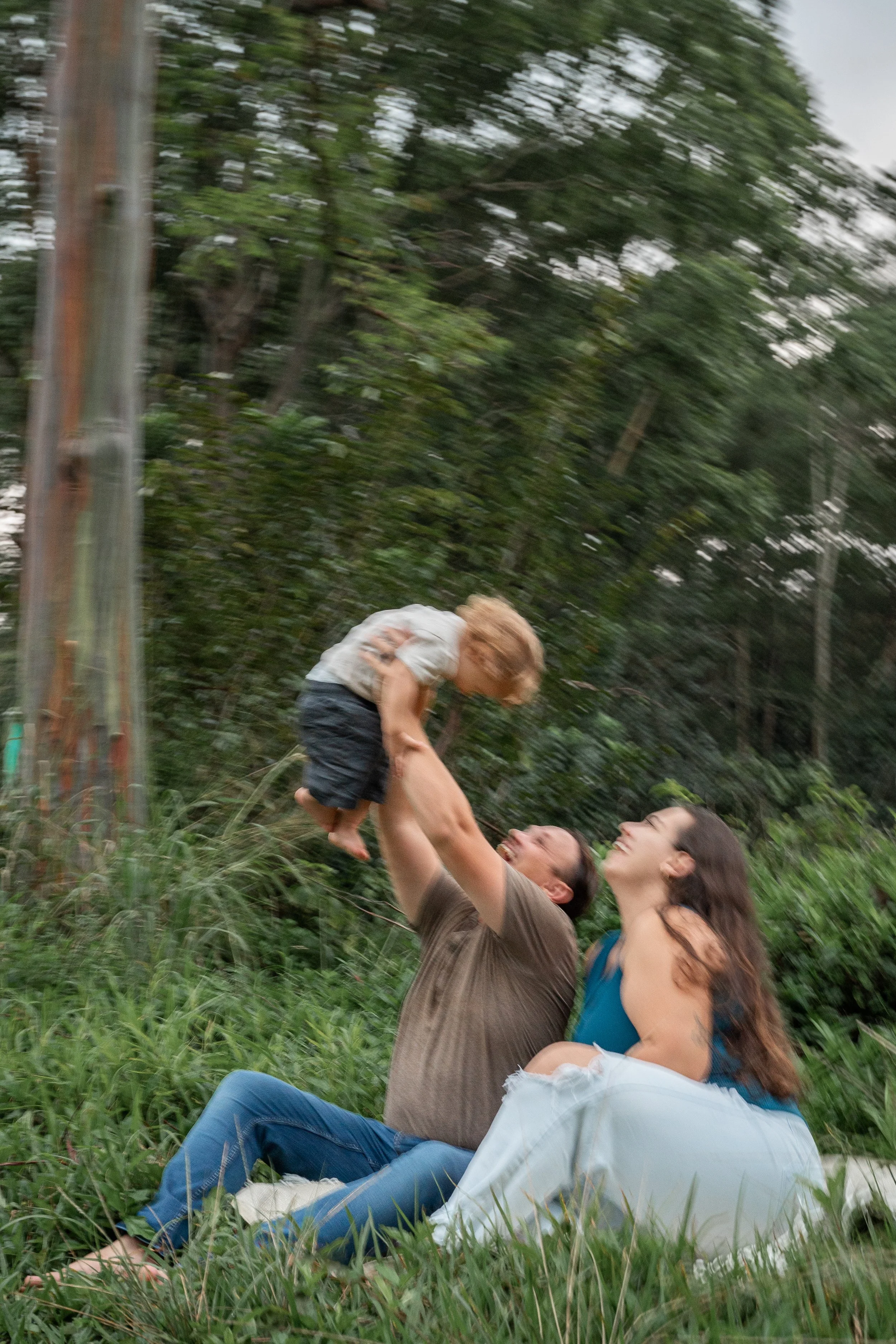 A family playing outdoors in a grassy, wooded area, with a man lifting a young child and both smiling. The child is reaching towards an adult woman sitting on the ground, surrounded by green trees and grass.